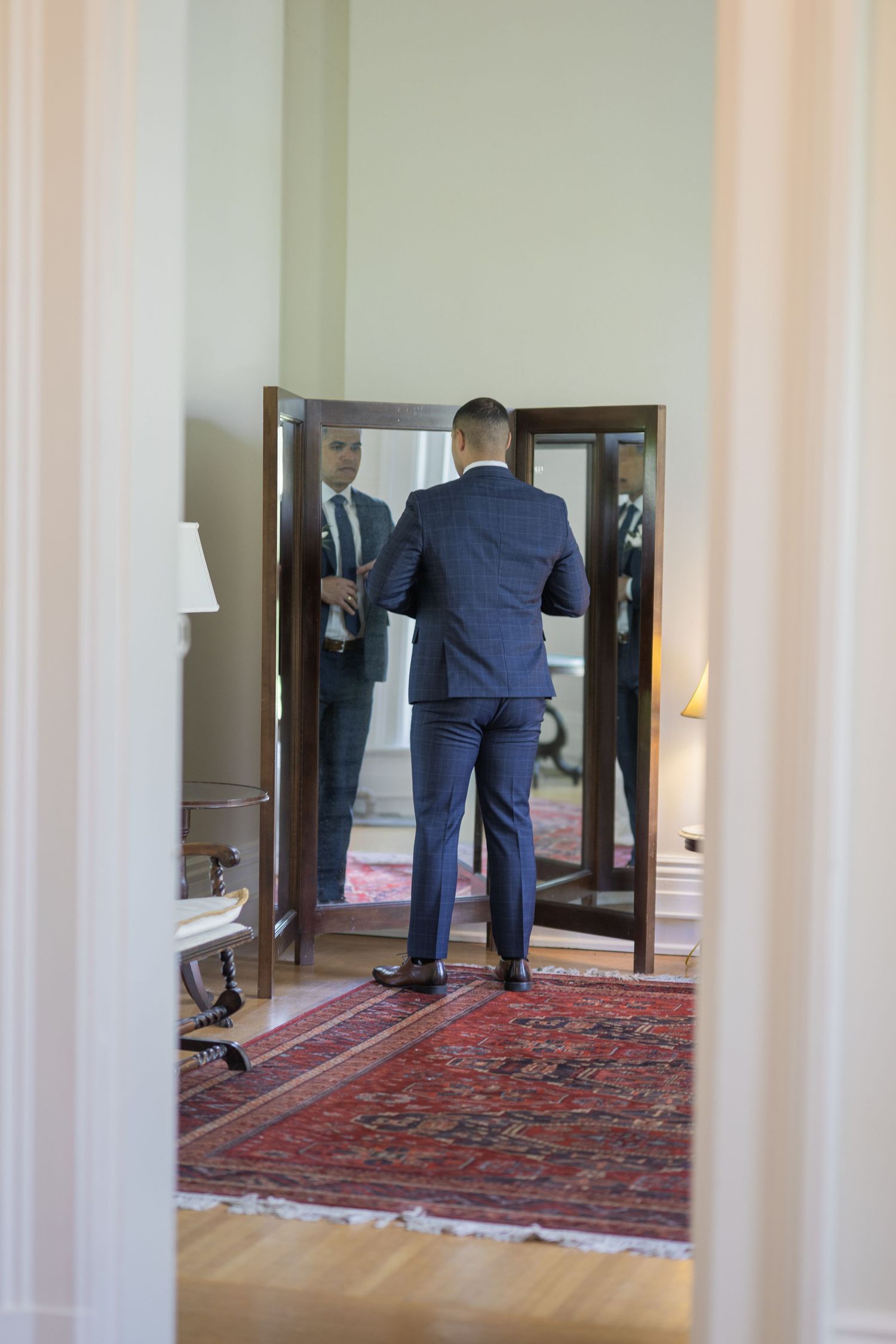 Groom in navy suit checking his appearance in a full-length mirror before wedding ceremony.