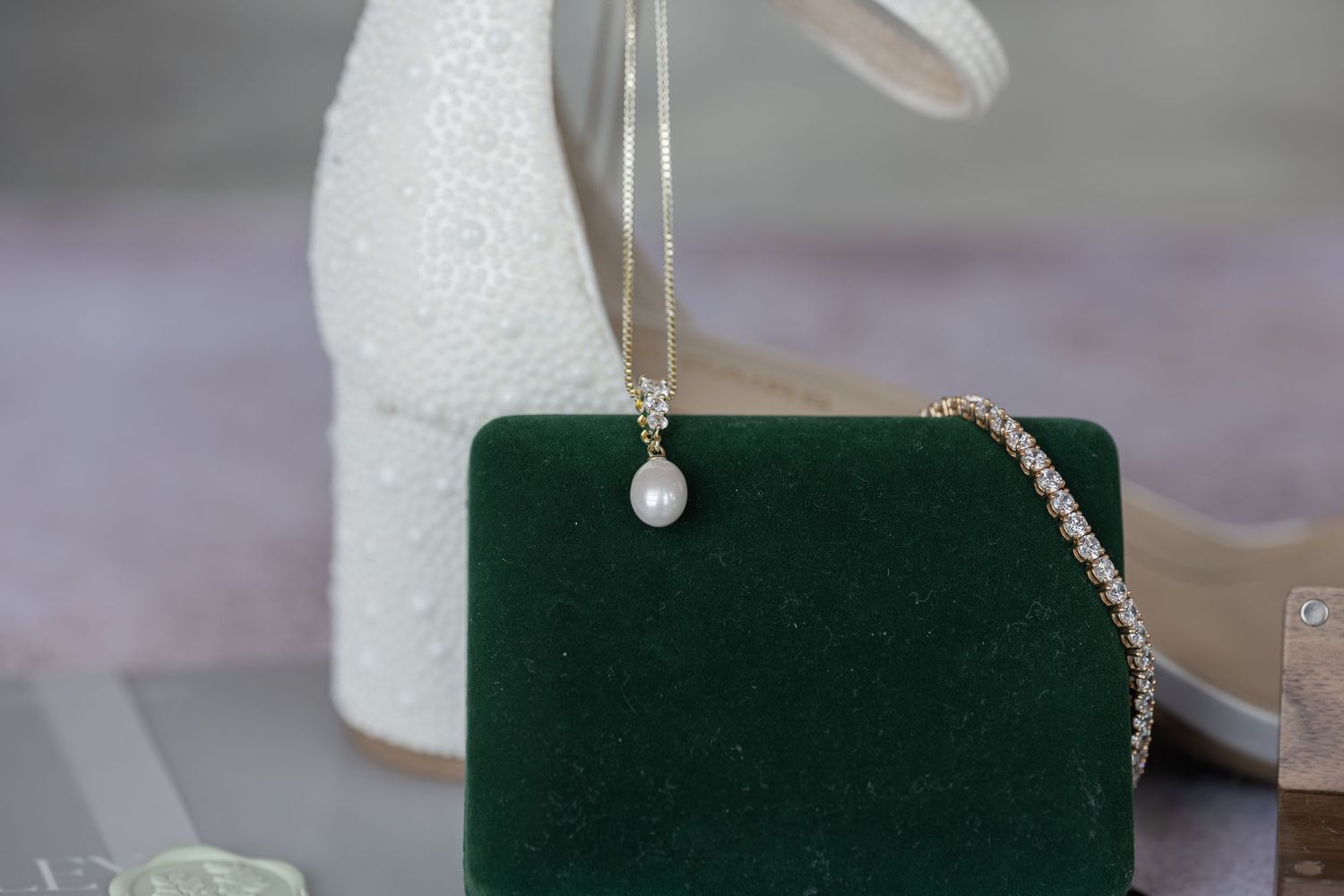 Elegant pearl drop necklace on green velvet display with white beaded bridal heels in background.