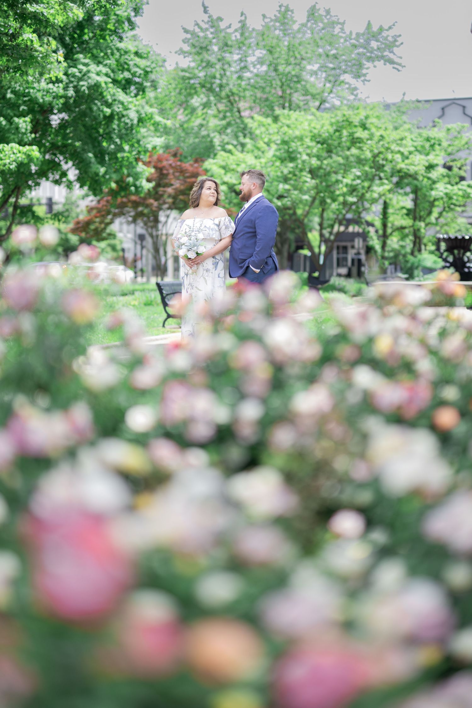 Couple standing among blooming pink roses in a lush green garden, woman in floral dress, man in blue suit.