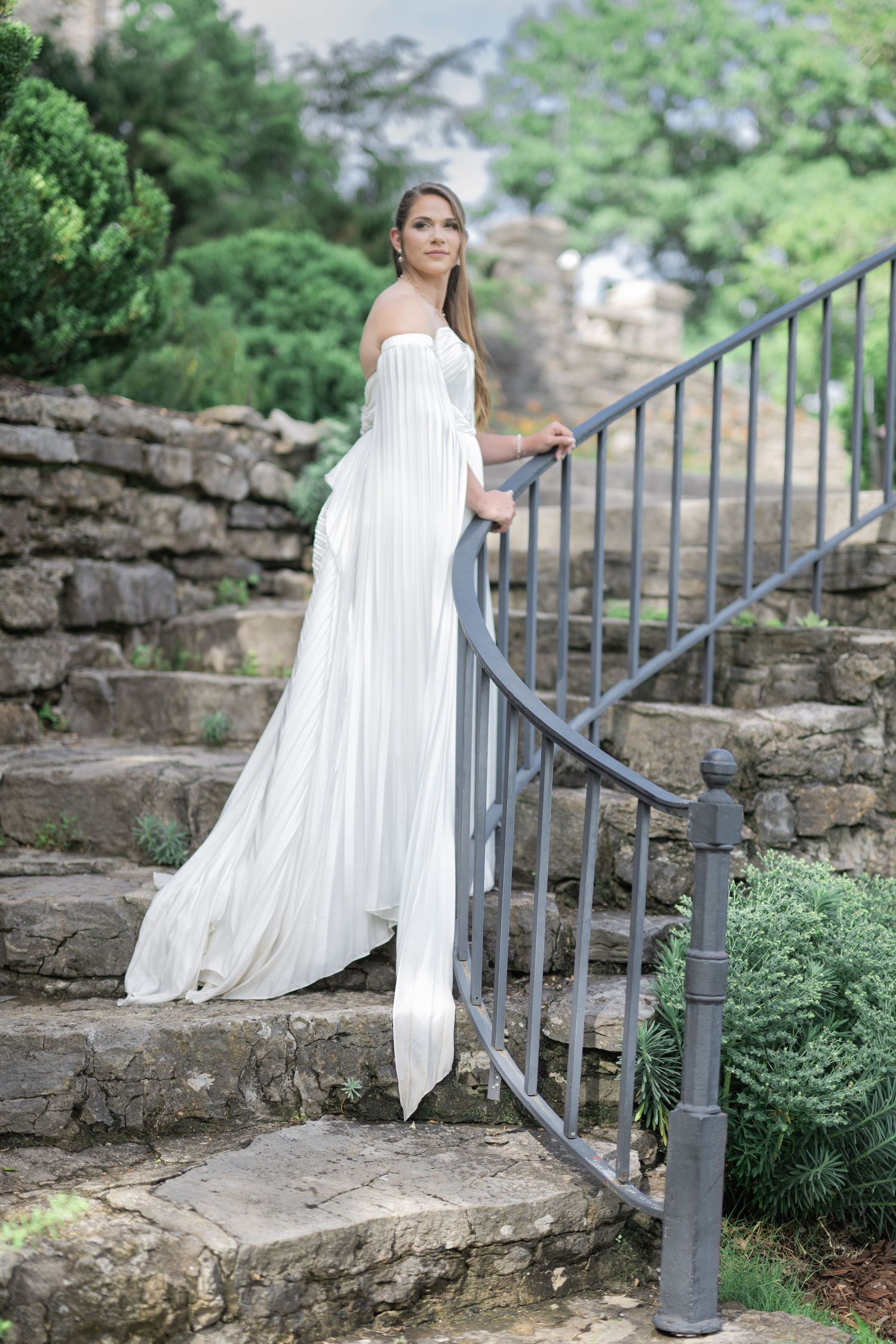 Elegant bride in flowing white off-shoulder gown standing on stone steps with iron railing surrounded by lush greenery.
