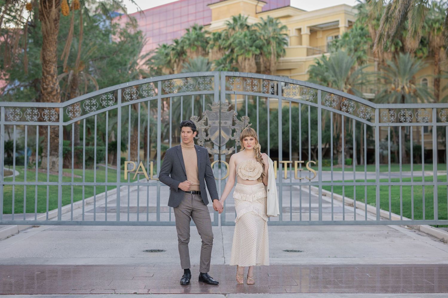 Couple poses in front of ornate iron gates surrounded by palm trees and lush greenery at an elegant venue.
