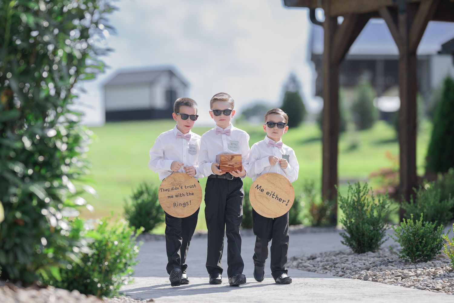 Three young ring bearers in suits and sunglasses holding round signs on a garden path at a wedding.