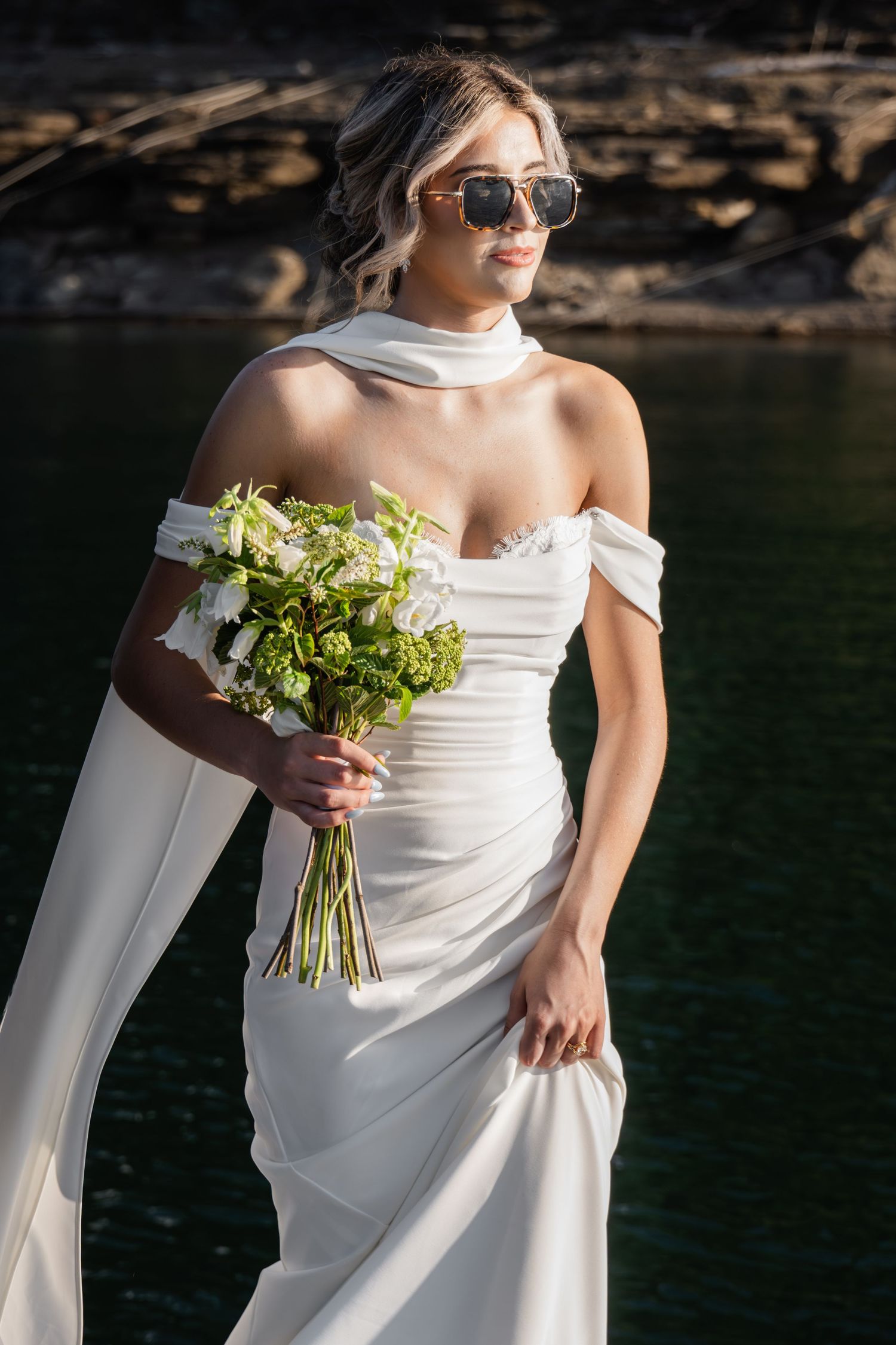 Bride in white off-shoulder gown and sunglasses holding green and white bouquet near rocky waterfront.