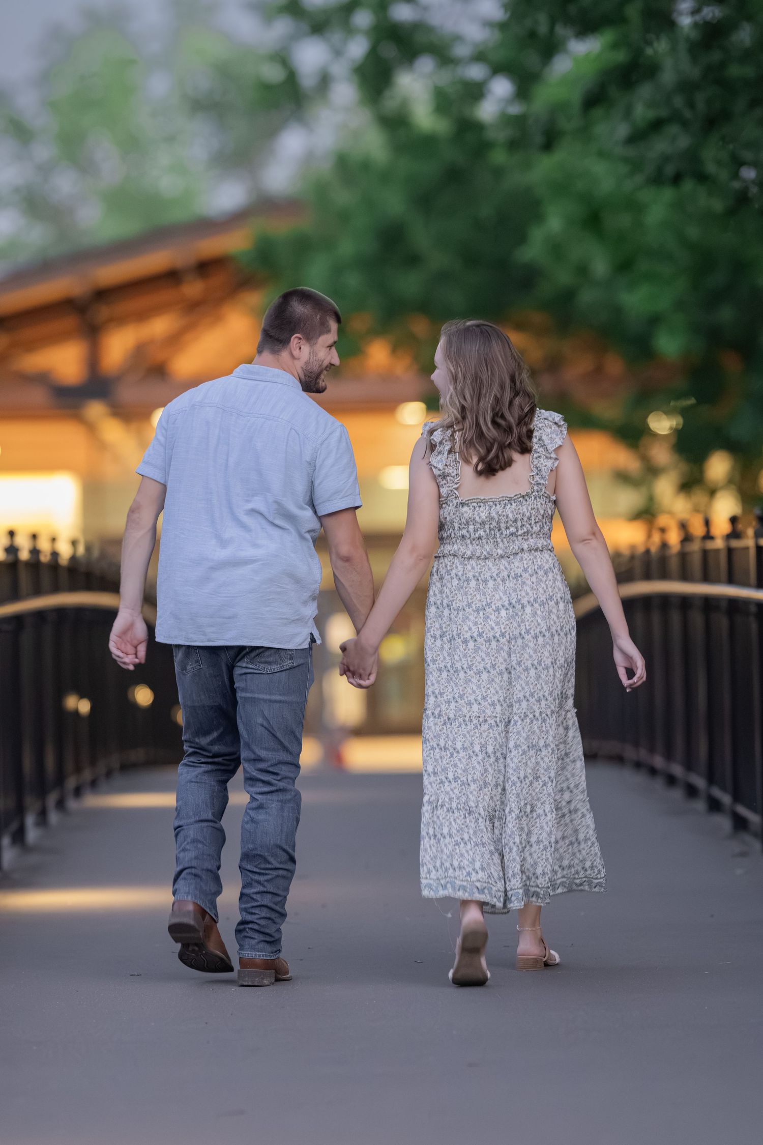 Couple holding hands walking away on a bridge path, man in blue shirt and jeans, woman in floral maxi dress.