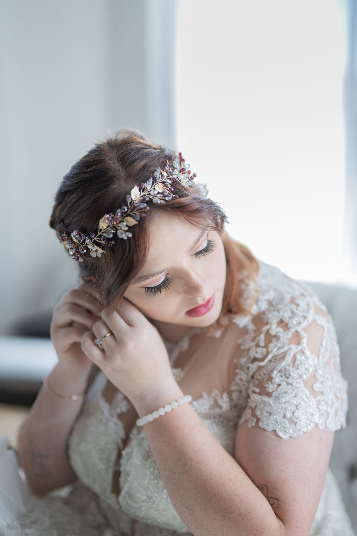 Bride in lace wedding dress wearing floral crown adjusts earring, looking down elegantly near window.