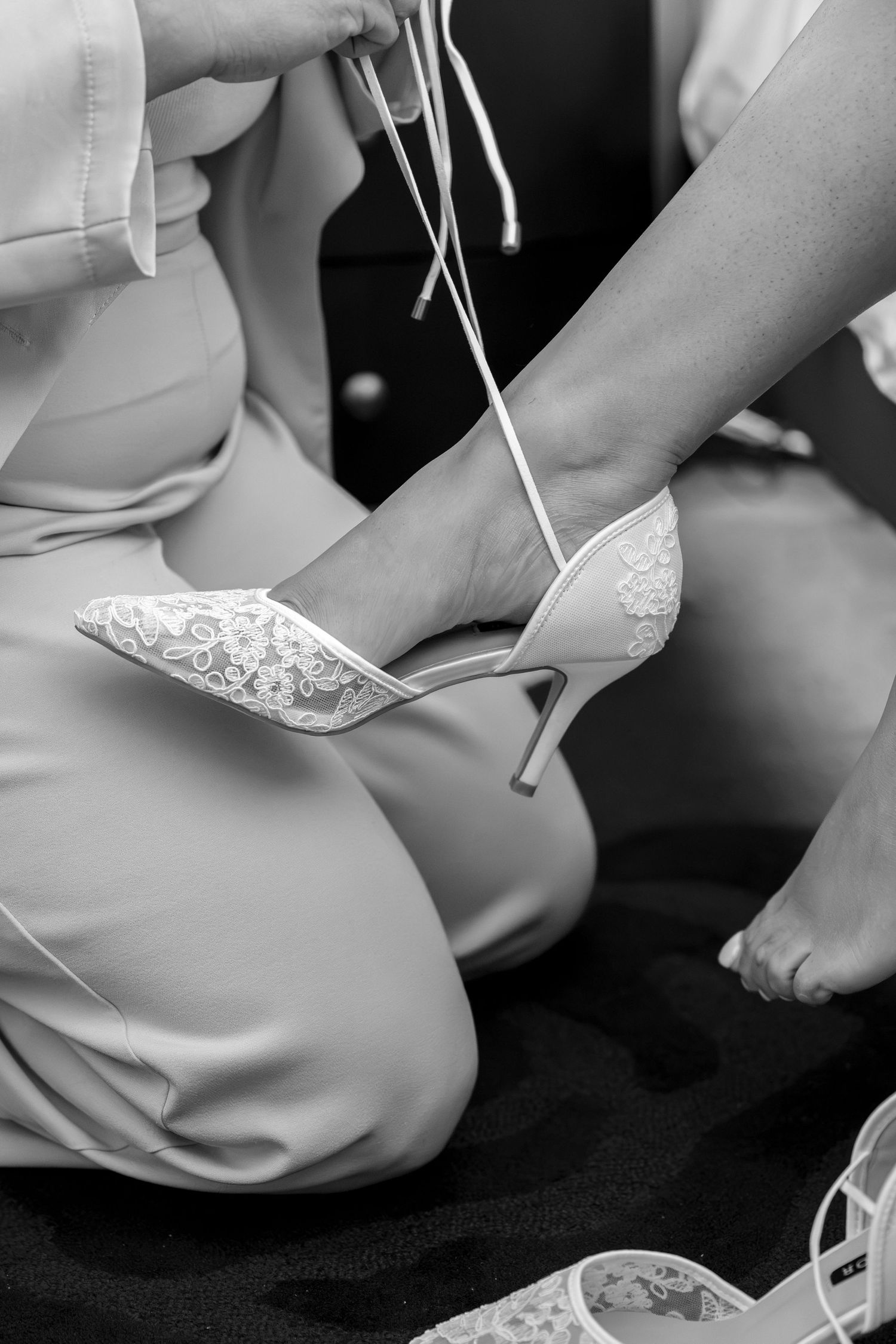 Bridesmaid helping bride put on white lace high heel wedding shoes in black and white photo.