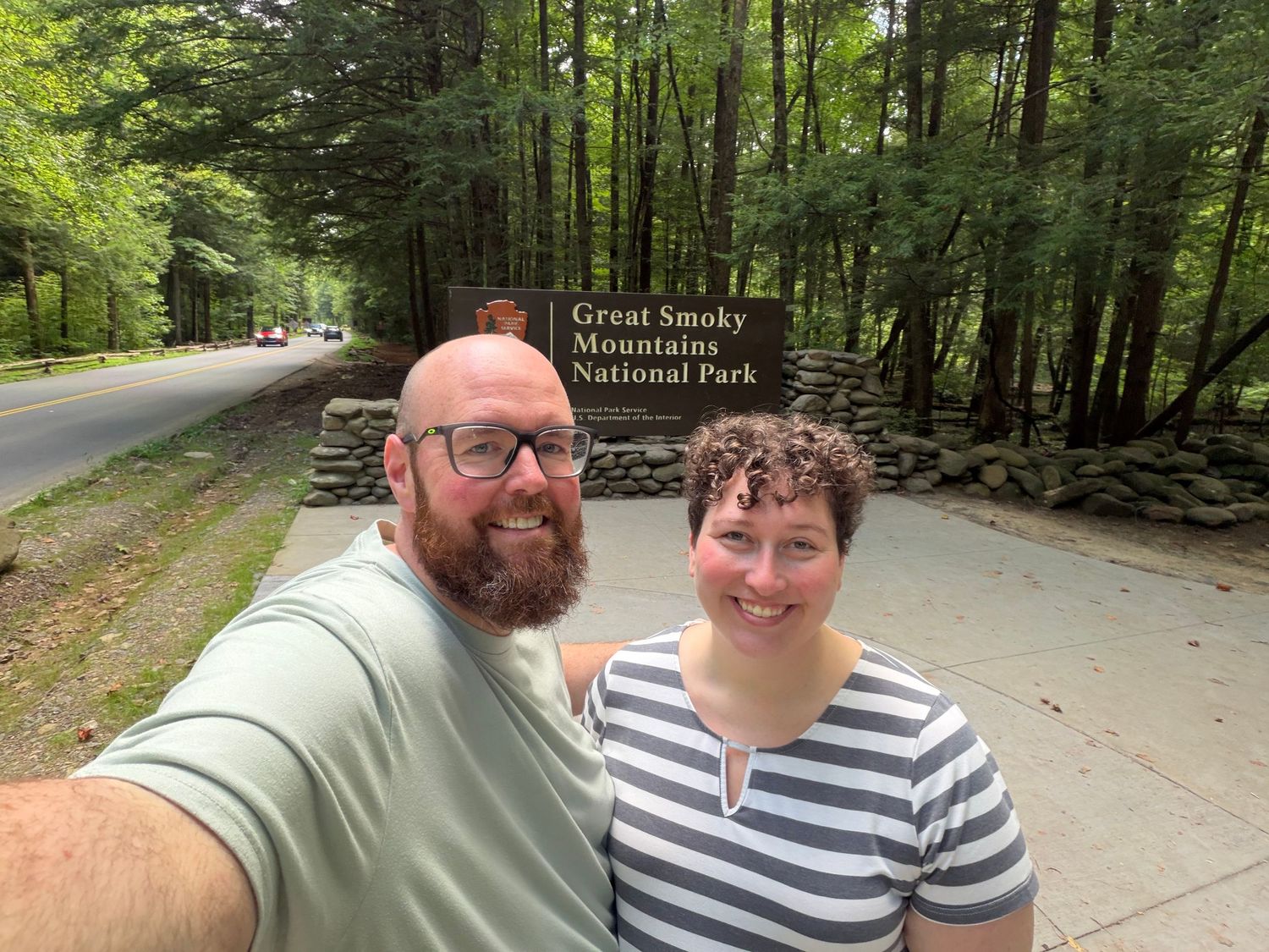 Couple takes selfie at Great Smoky Mountains National Park entrance sign surrounded by lush green forest trees.