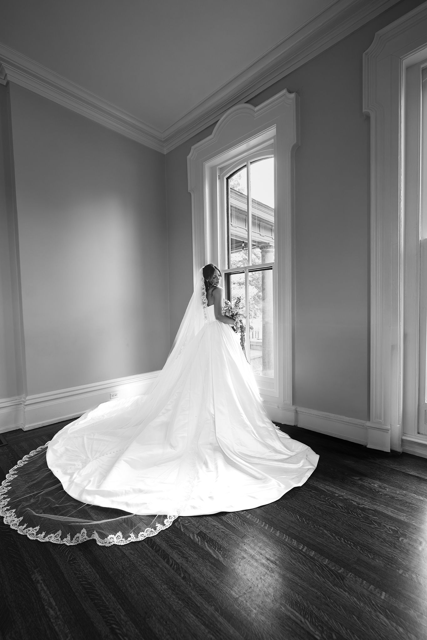 Bride in elegant white gown with long cathedral veil stands by window in black and white bridal portrait.