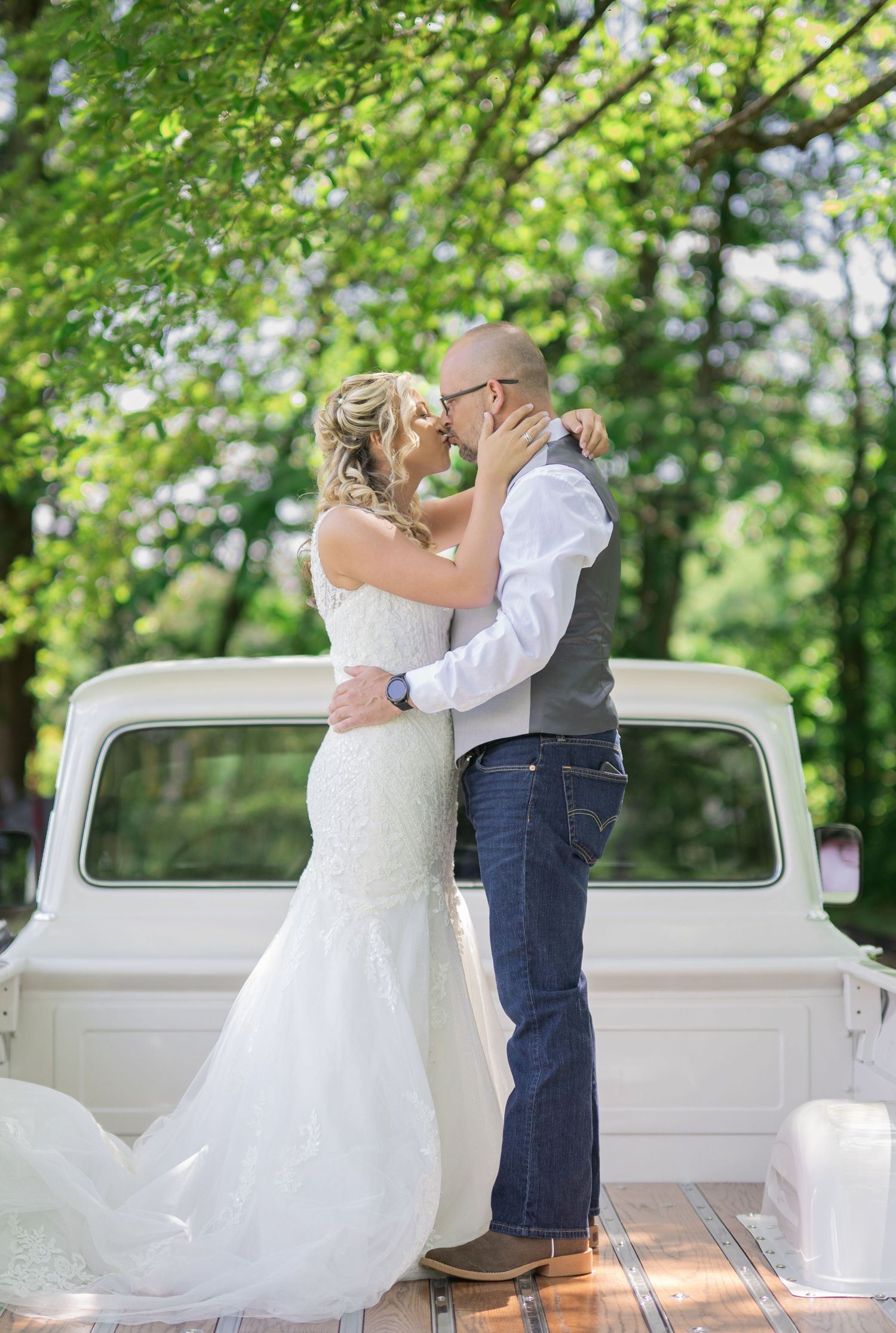 Bride and groom sharing a kiss in front of a vintage white truck surrounded by lush green trees on their wedding day.