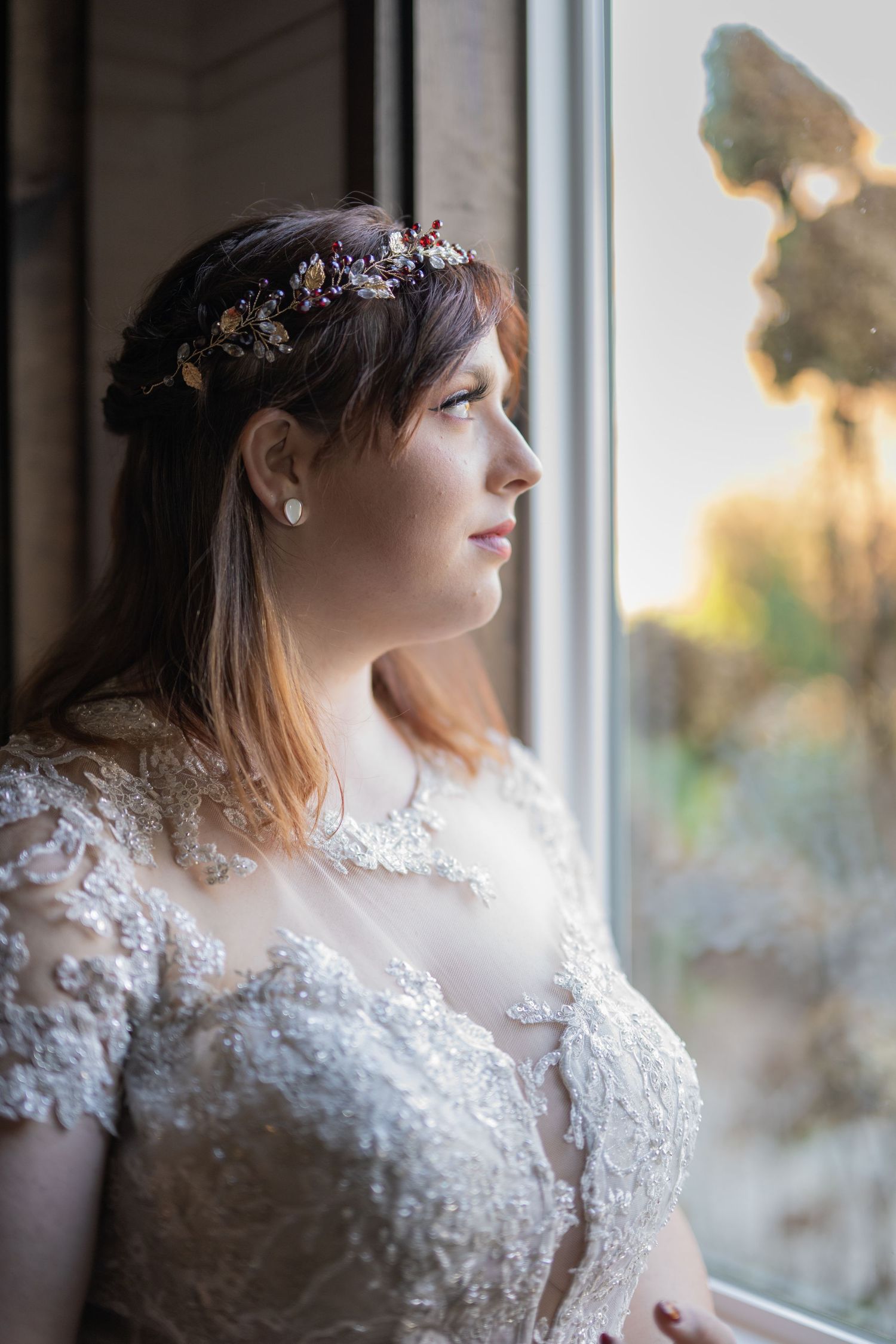 Bride in lace gown and floral crown gazes thoughtfully out a sunlit window on her wedding day.