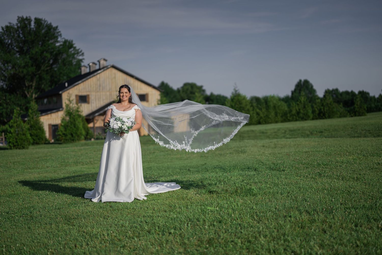 Bride in white gown with flowing veil stands on green lawn before rustic barn venue on a clear day.