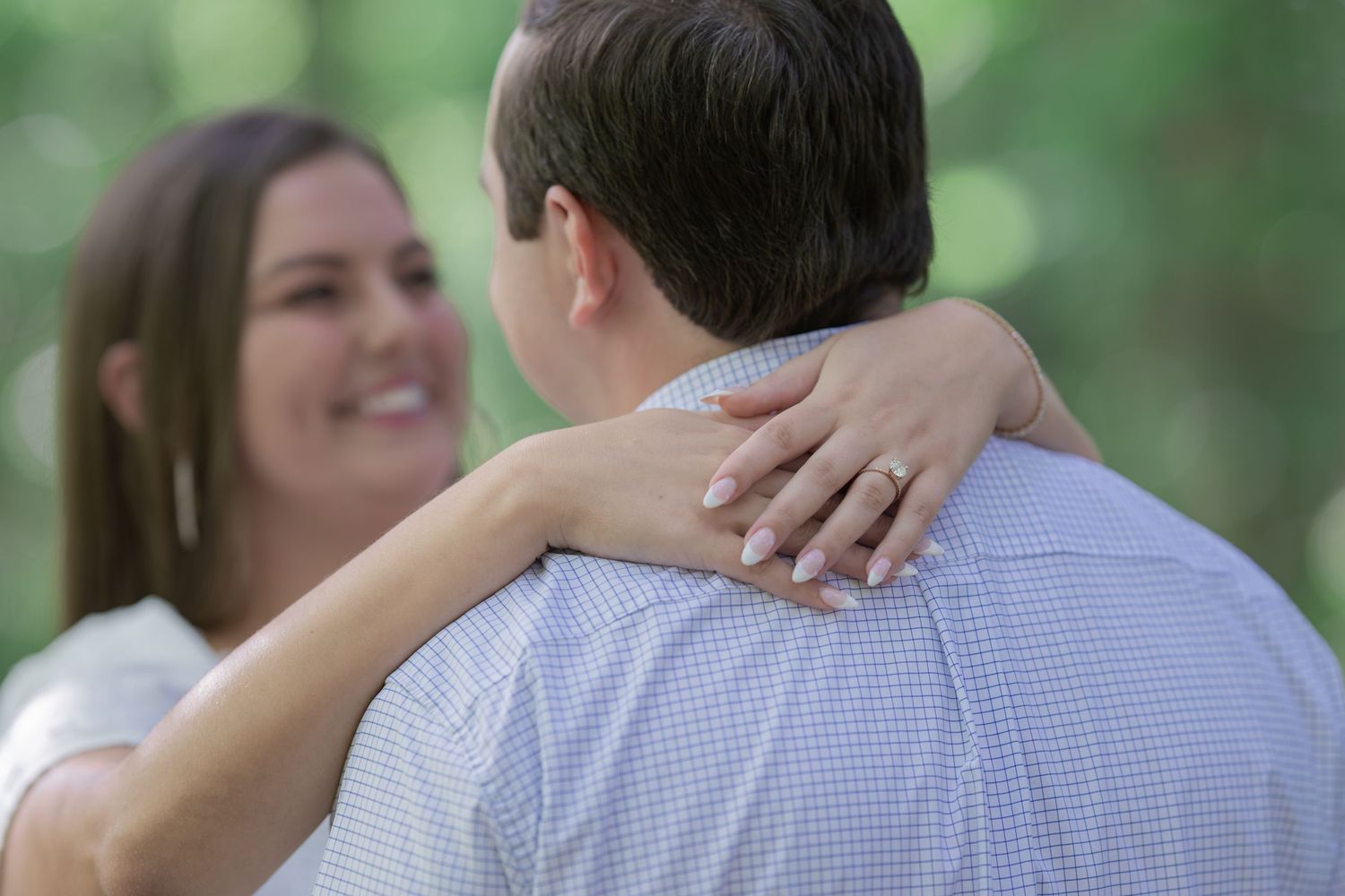 Couple embracing outdoors, woman smiling with engagement ring visible on hand resting on man's shoulder.