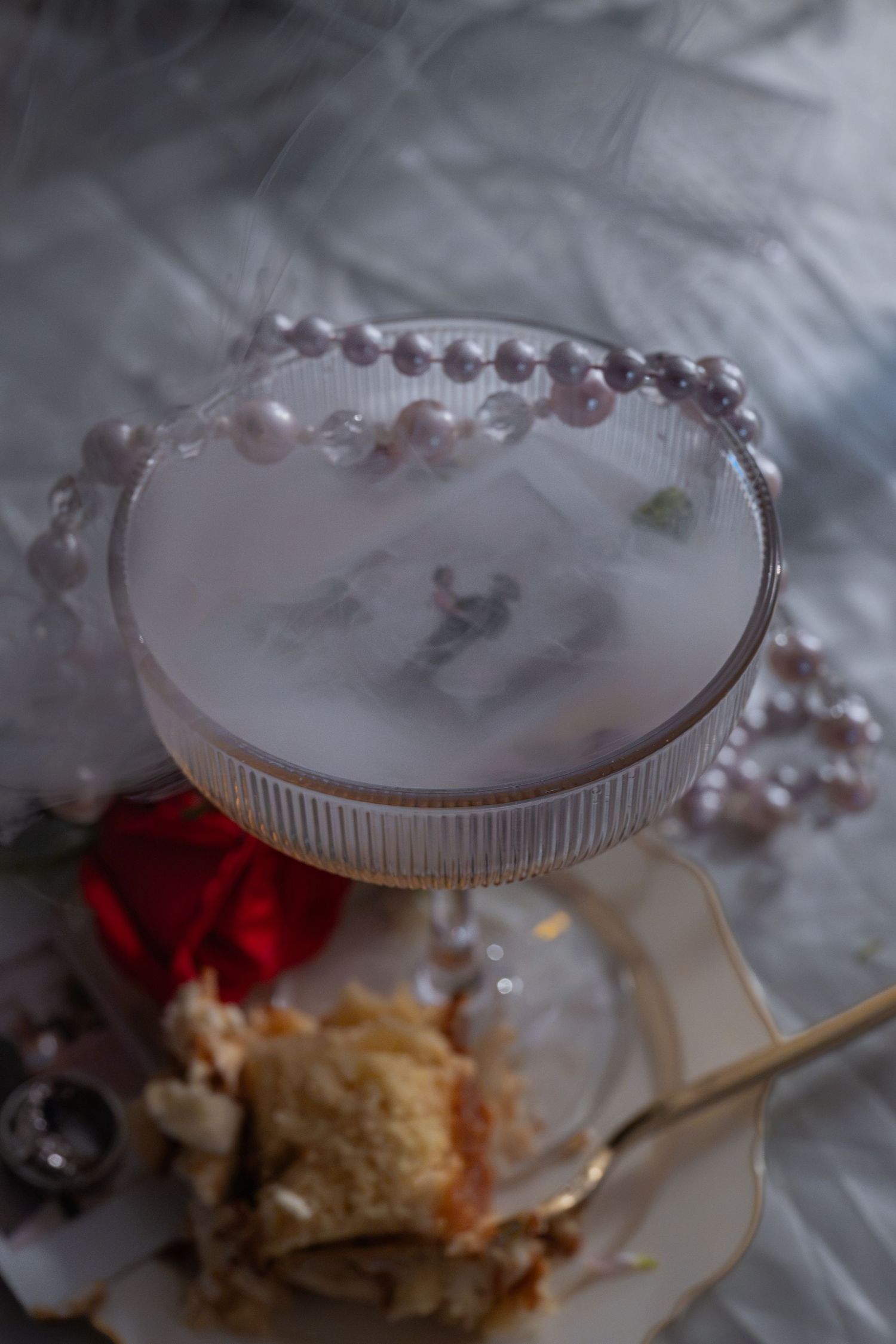 Elegant cocktail in ribbed coupe glass with pearl necklace, dry ice fog, beside red rose and cake slice.