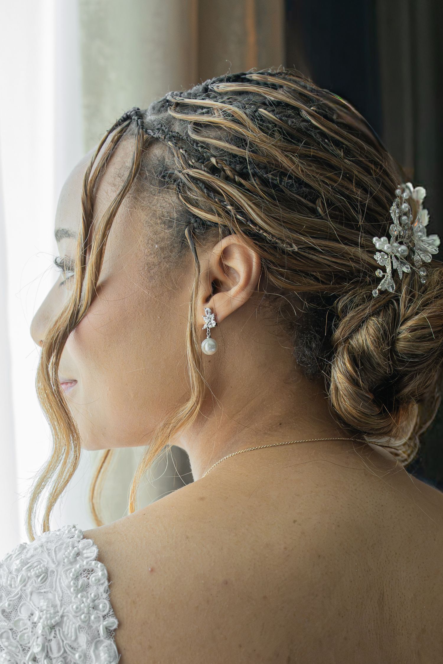 Bride with braided updo, pearl drop earrings, and floral hair accessory, wearing a lace wedding dress.