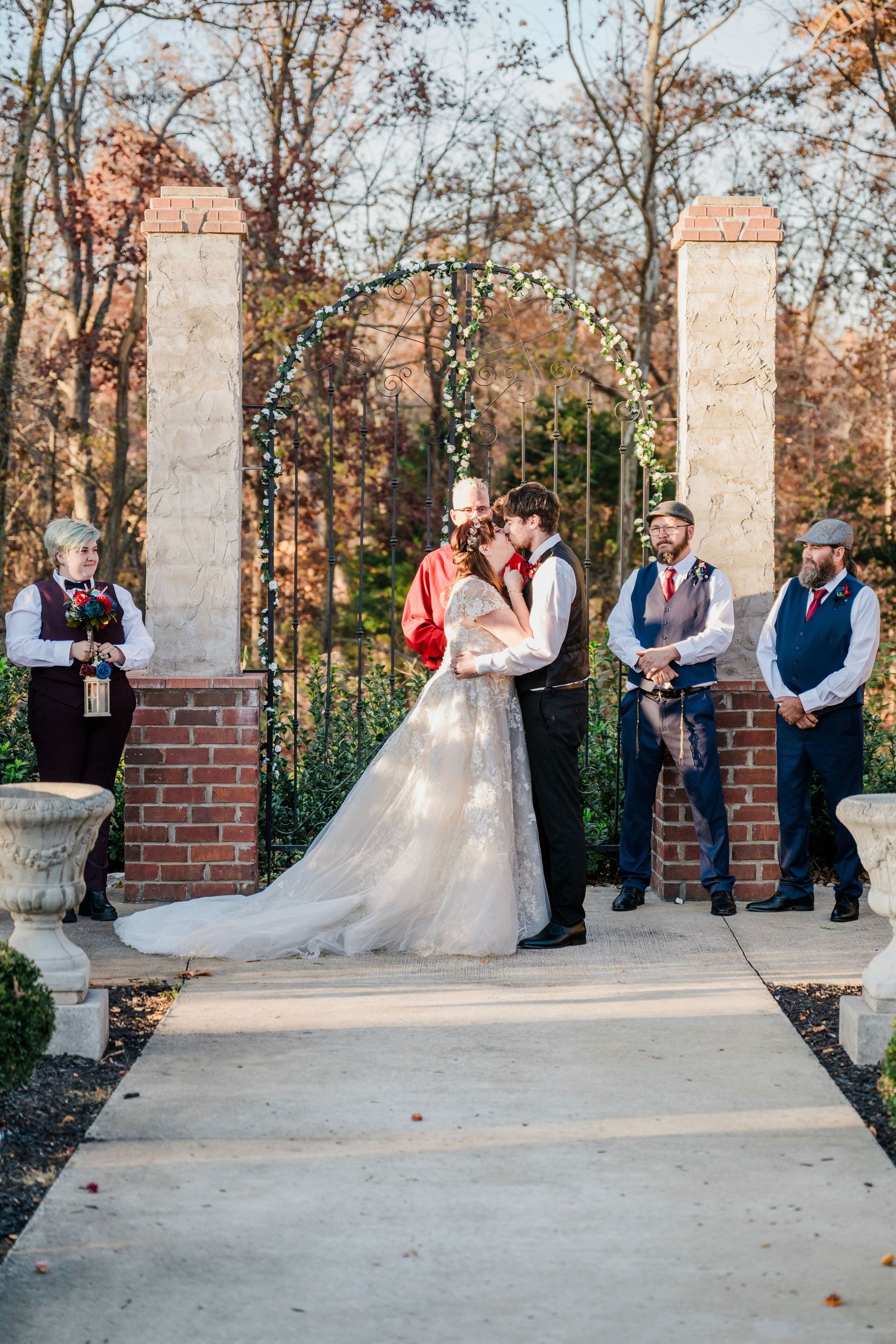Bride and groom share a kiss at outdoor wedding ceremony with arch, groomsmen, and officiant in autumn garden setting.