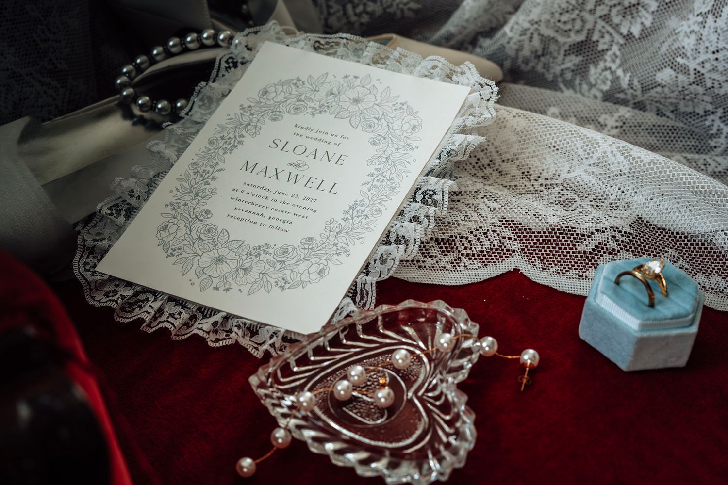 Elegant wedding invitation with floral border beside pearl jewelry, diamond ring, and earrings on red velvet.