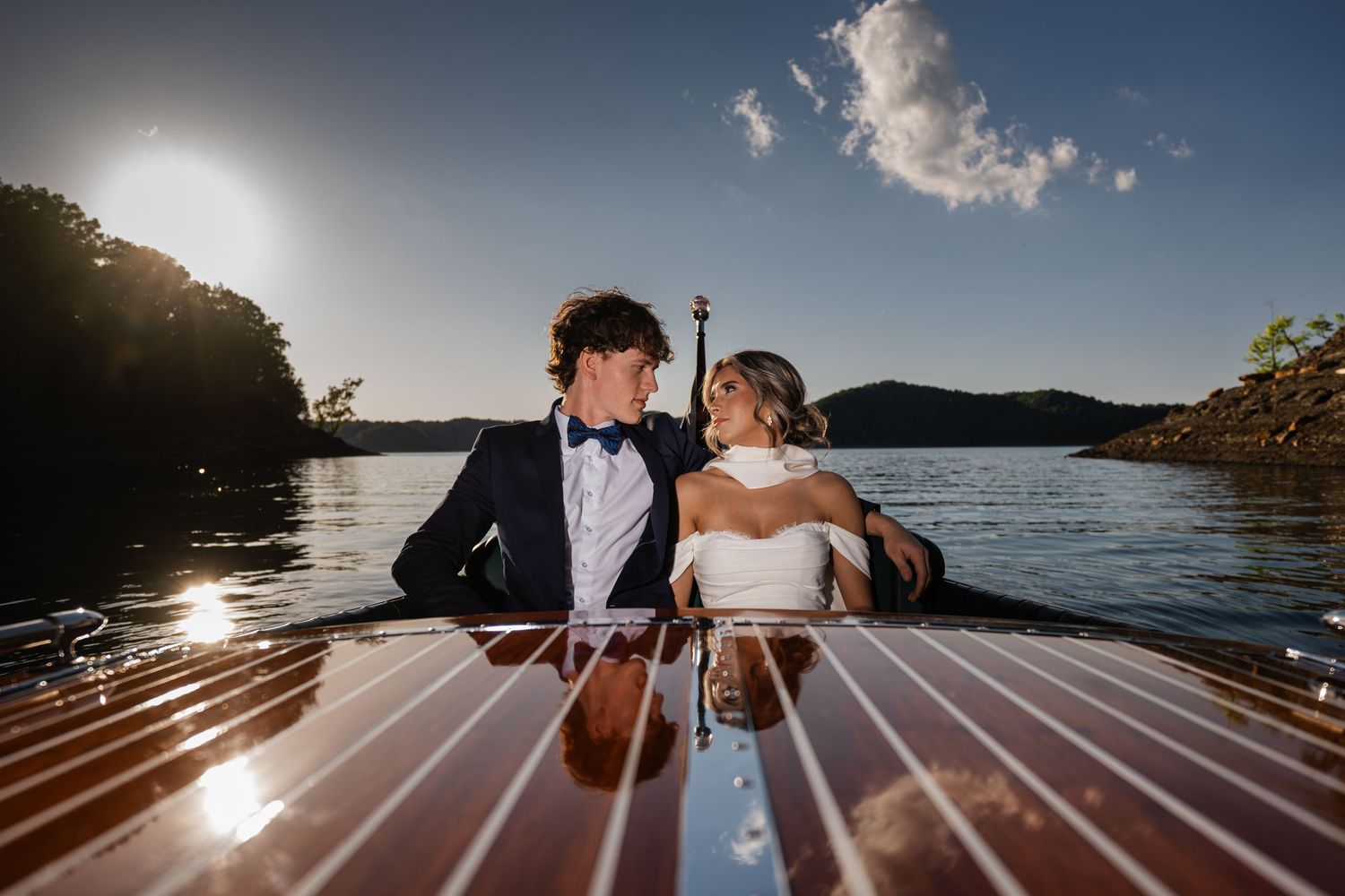 Bride and groom sitting together on a wooden boat on a sunlit lake, sharing a romantic moment on their wedding day.