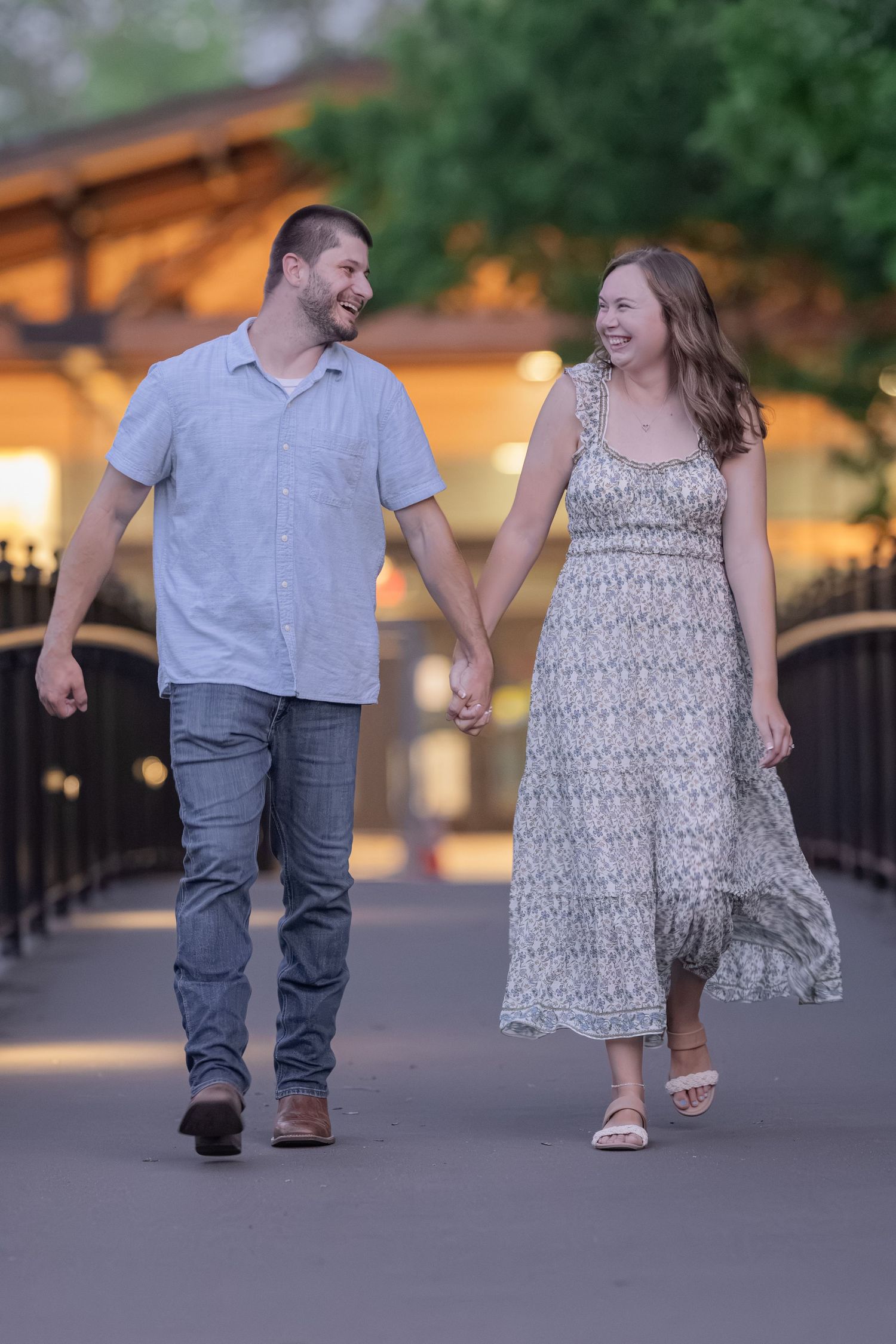 A smiling couple holds hands and walks together on a bridge path at dusk, man in blue shirt and woman in floral dress.