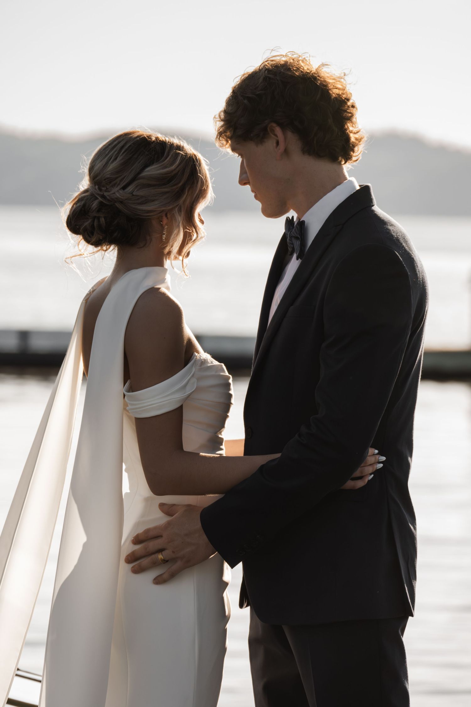 Elegant bride and groom share an intimate moment by the water, wearing a white gown and black tuxedo.