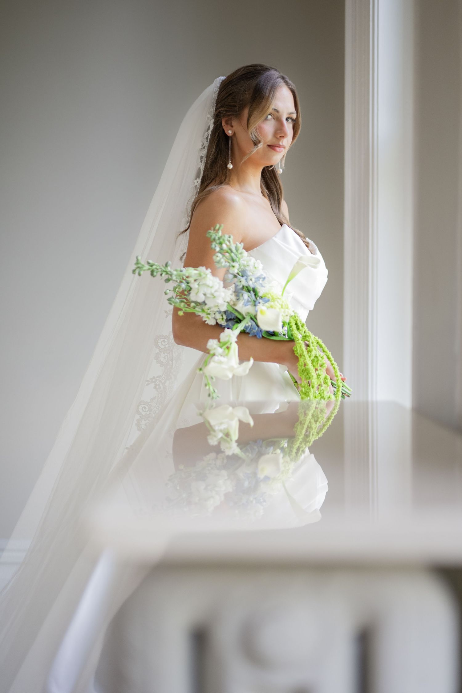 Bride in white off-shoulder gown holds floral bouquet with green accents, wearing a long veil near a bright window.
