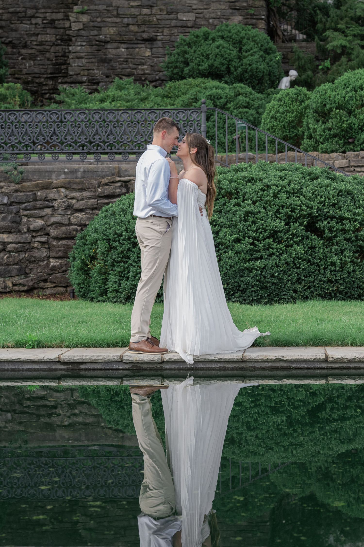 Bride and groom sharing a romantic moment by a reflective pool in a lush garden setting.