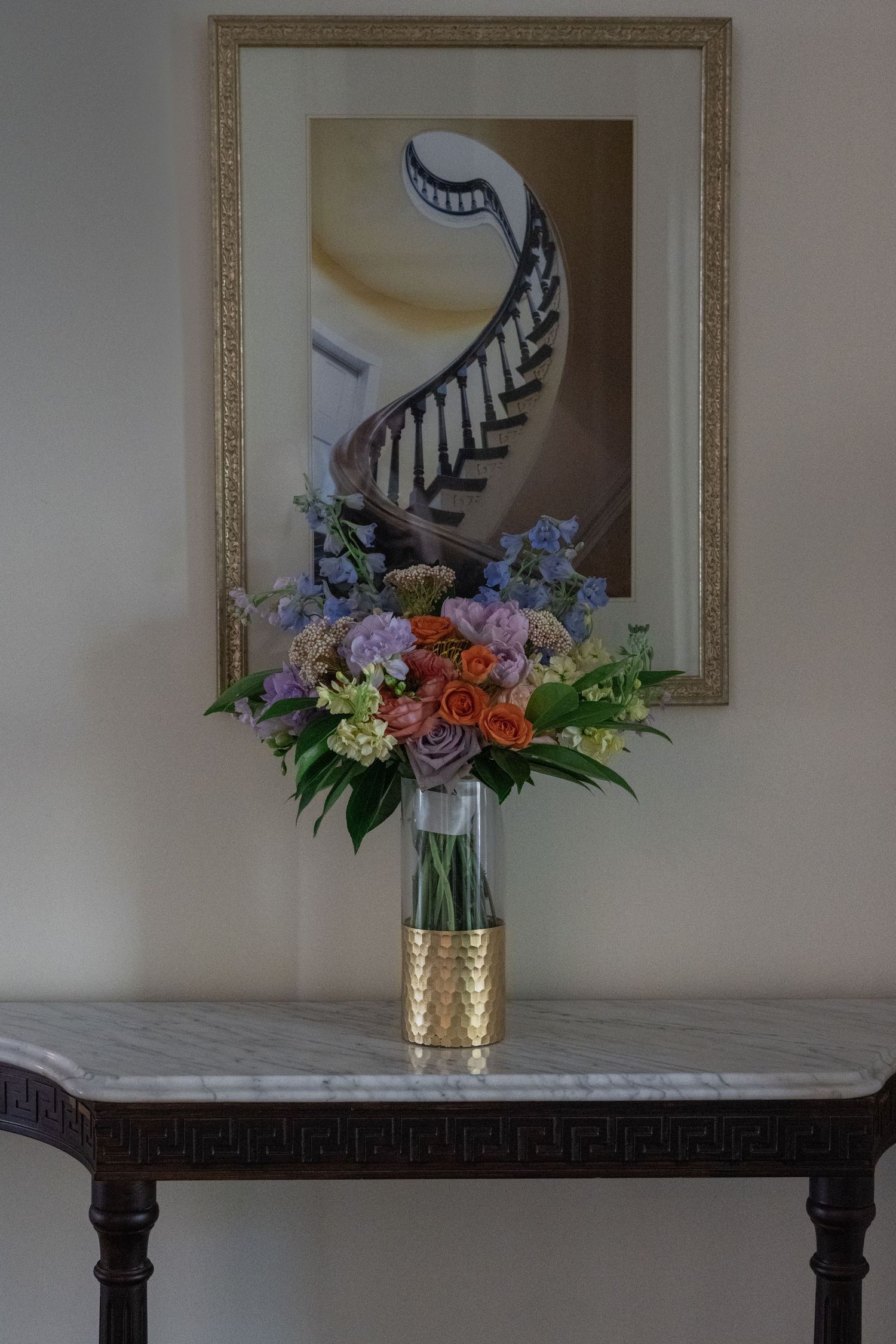 Colorful floral bouquet in gold vase on marble table with framed spiral staircase artwork on wall.