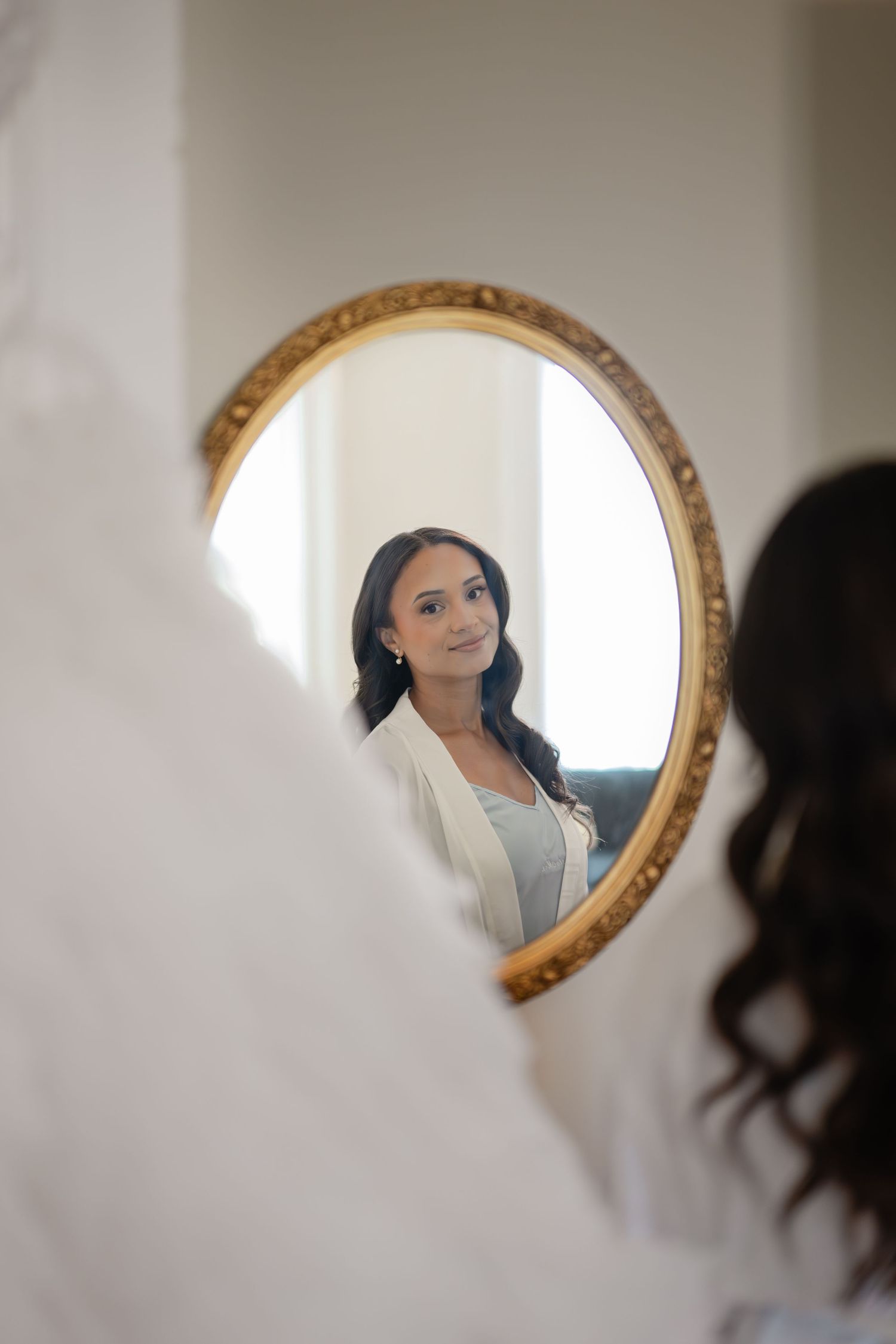A woman with dark hair gazes at her reflection in an ornate gold oval mirror while holding a white wedding dress.