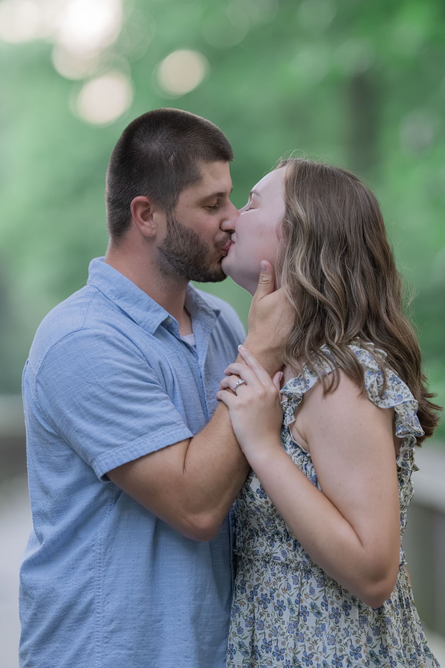 A couple shares a romantic kiss outdoors, the woman in a floral dress and man in a blue shirt, surrounded by lush greenery.