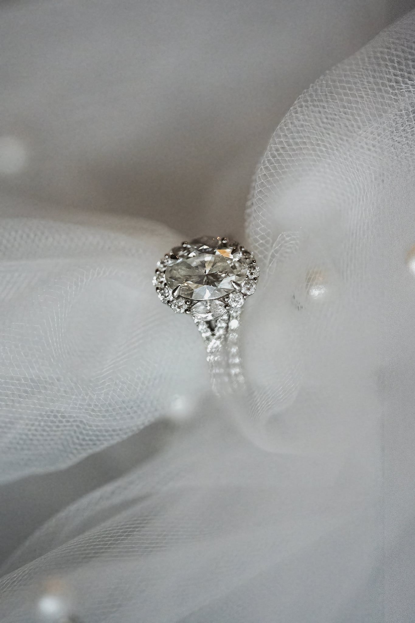 Elegant oval diamond halo engagement ring with twisted band resting on white bridal veil with pearls.