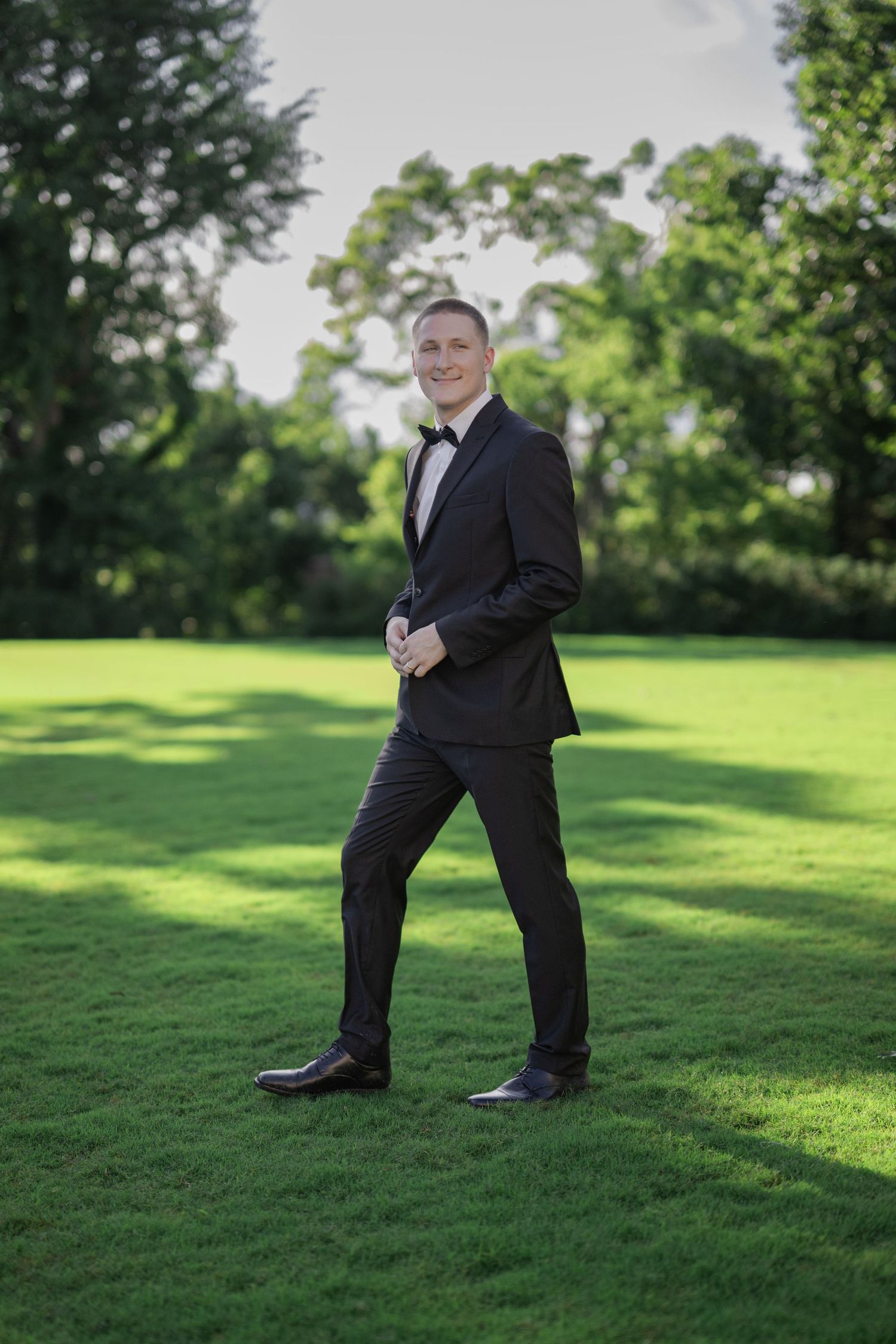 A man in a black tuxedo with bow tie stands smiling on lush green grass with trees in the background.