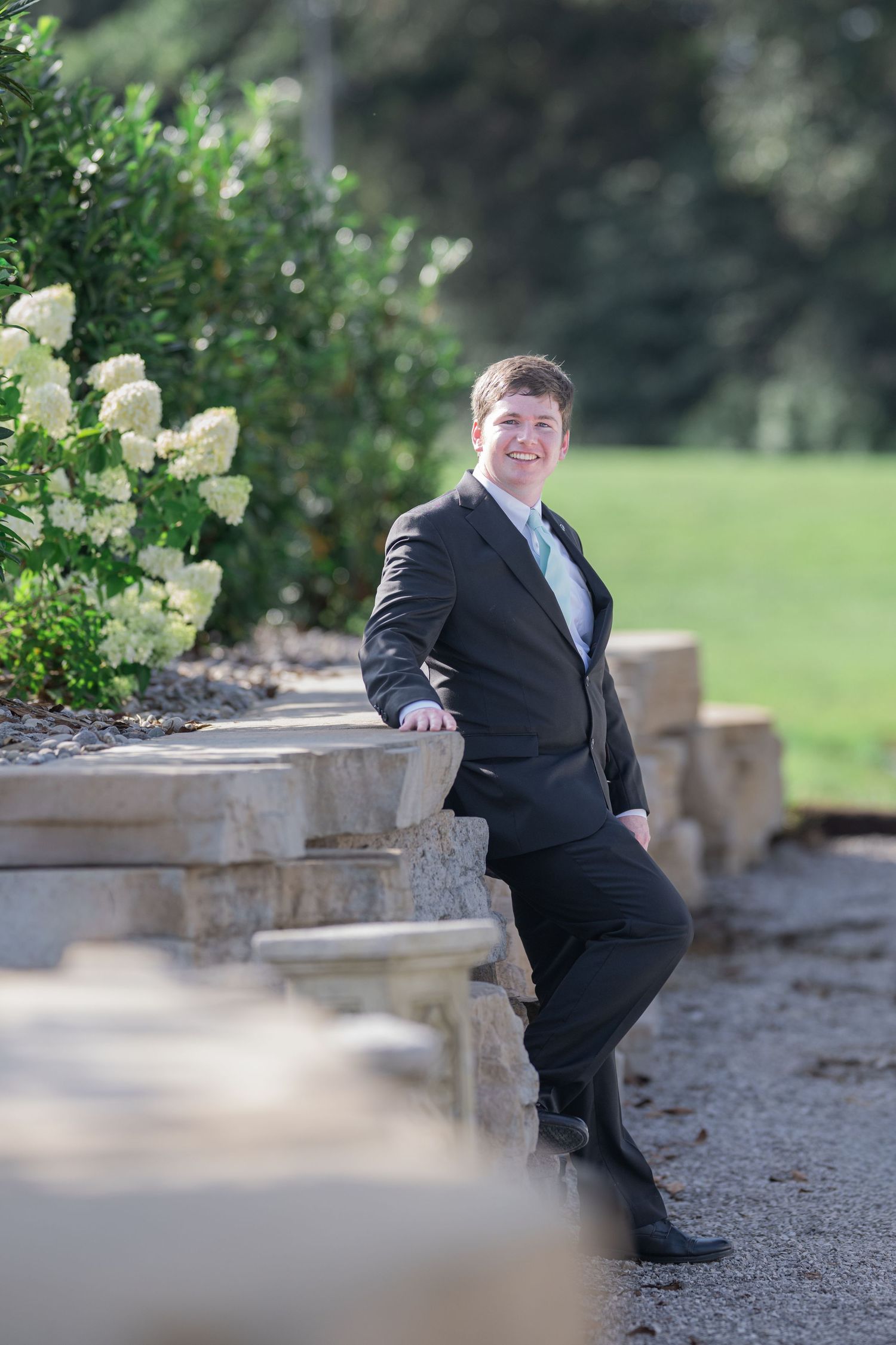 Young man in black suit leaning against stone wall in garden with white hydrangeas, smiling outdoors.