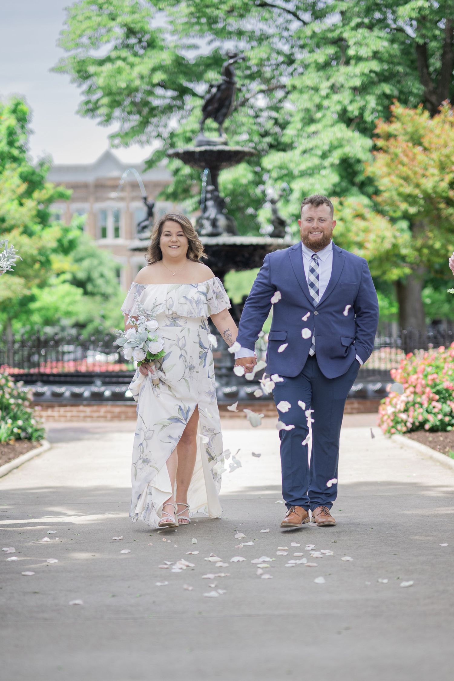 Bride and groom walk hand-in-hand through a park with a fountain, she holds white flowers, he wears a navy suit.