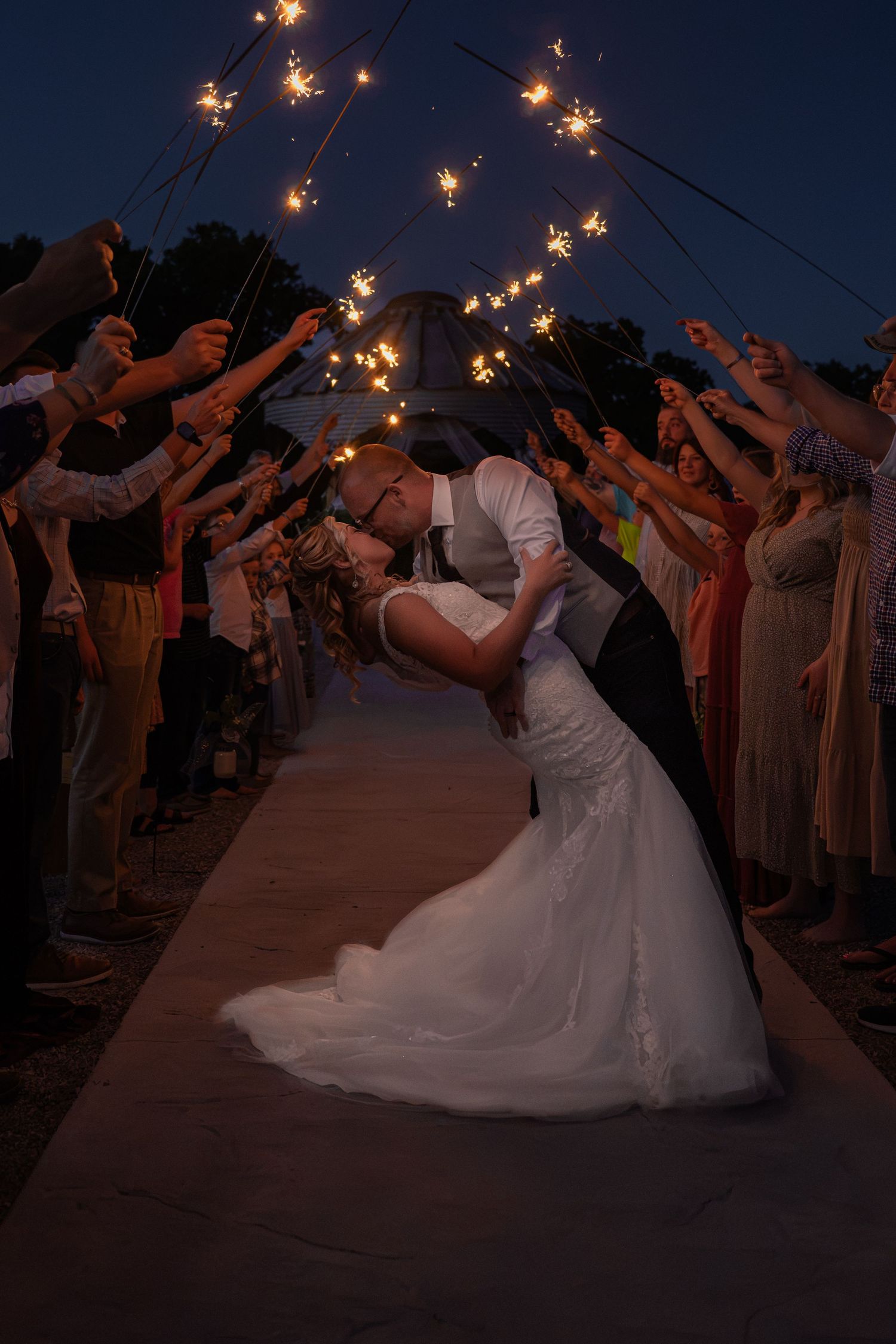 Bride and groom share a romantic dip kiss under a sparkling archway of wedding sparklers held by guests at night.