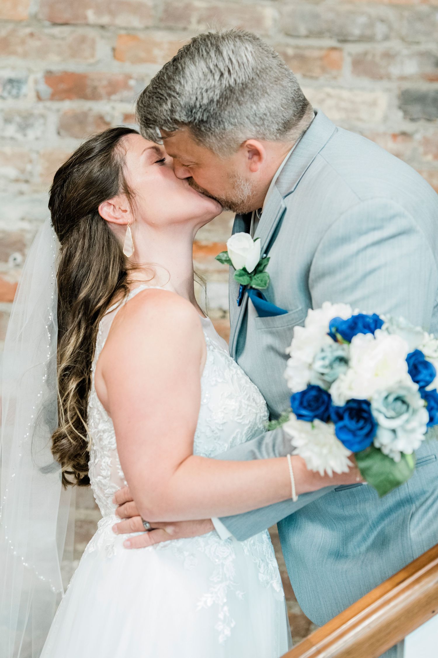 Bride and groom share a kiss at their wedding, with a blue and white bouquet, brick wall background.