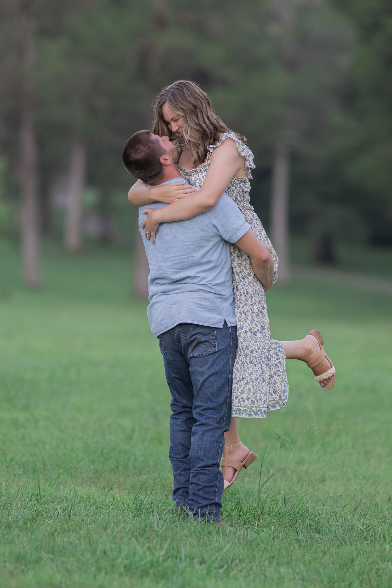 A man lifts a woman in a floral dress as they embrace and smile in a lush green park setting.