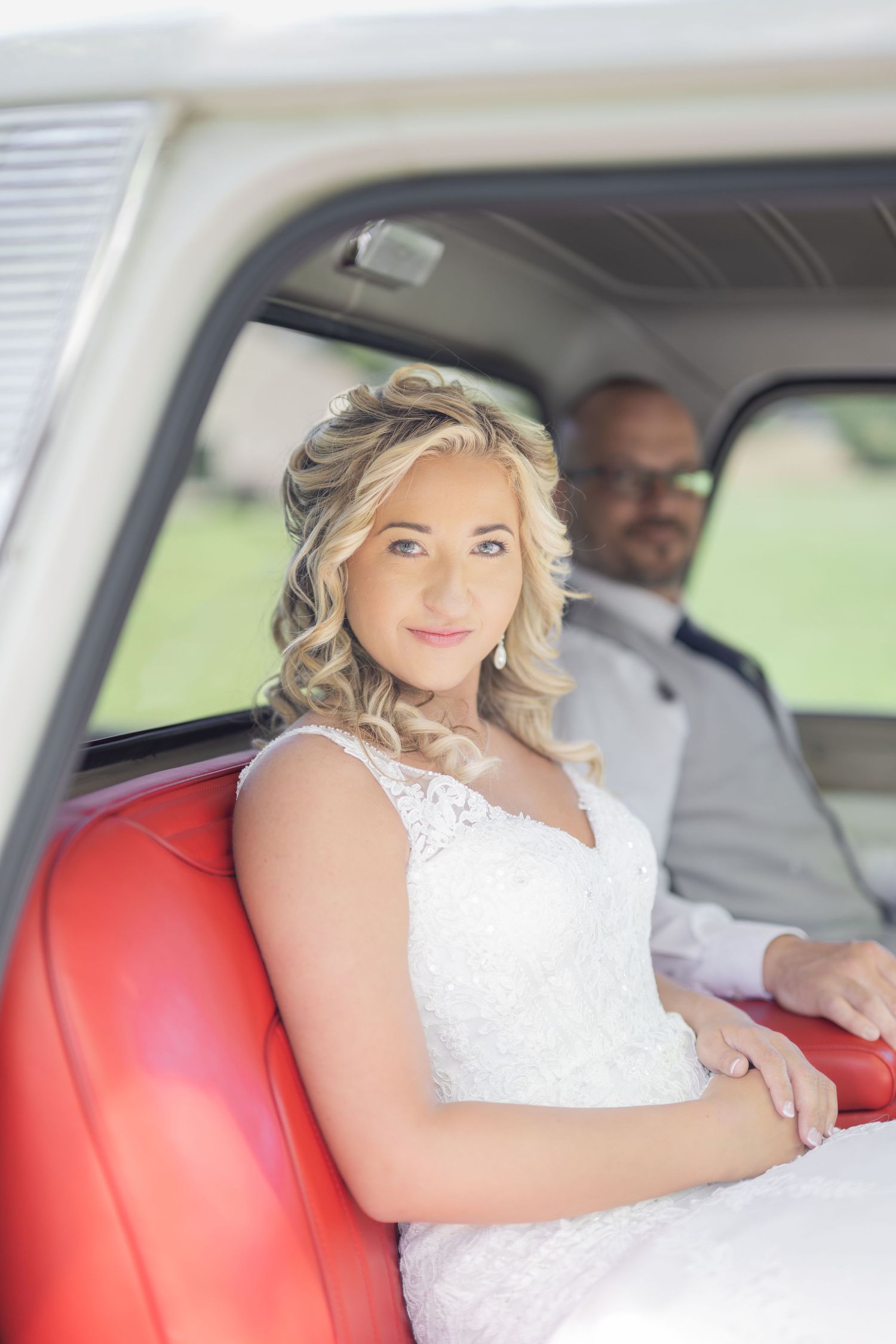 Bride in white lace dress sits on red leather seat in vintage vehicle, groom visible behind her.