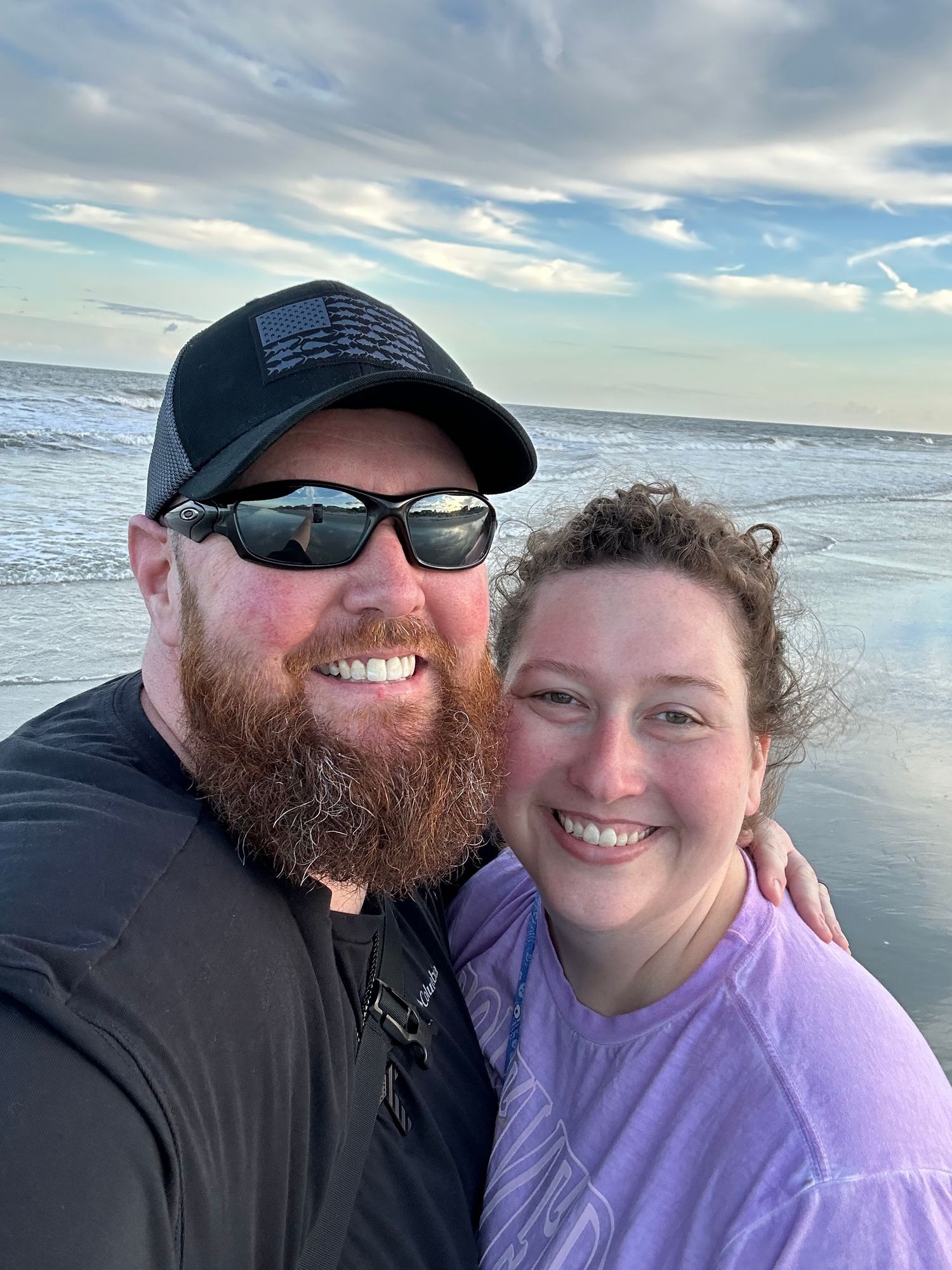 Smiling couple taking a selfie on a beach at sunset, man with red beard and black cap, woman in purple shirt.