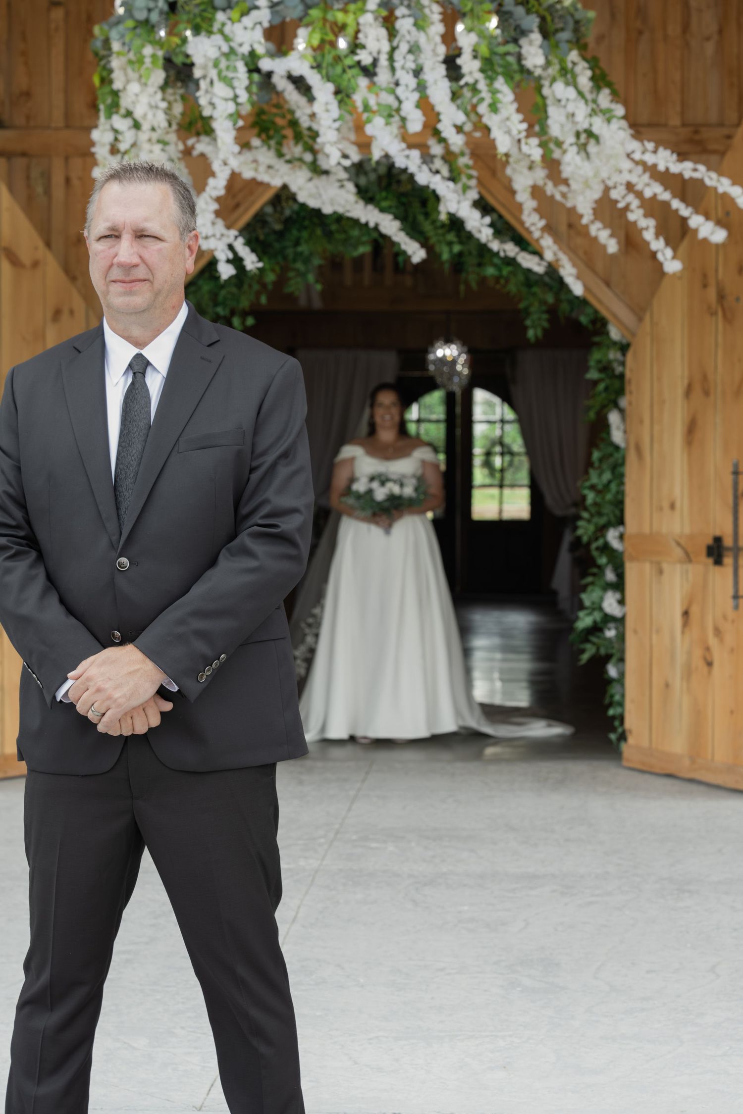Groom in dark suit stands waiting as bride approaches in decorated barn venue.