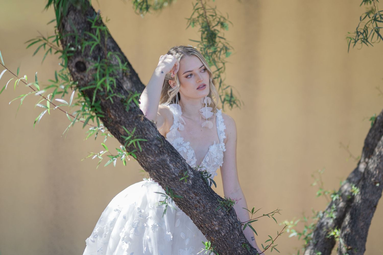 A bride in a white floral gown poses gracefully among tree branches with green leaves and a soft beige background.