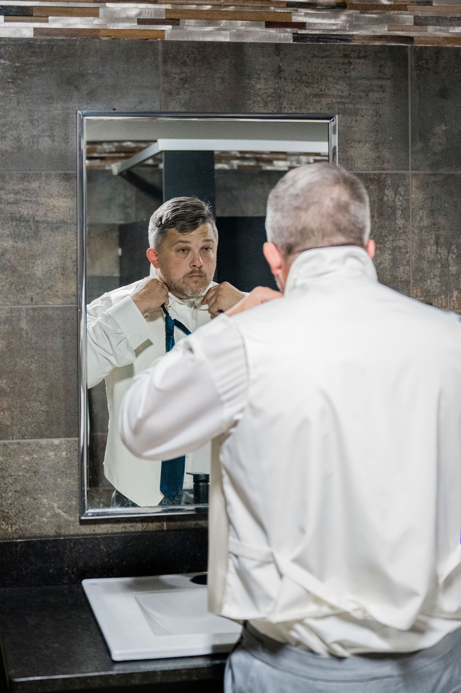 Man in white dress shirt adjusting tie while looking in bathroom mirror, getting dressed for formal occasion.
