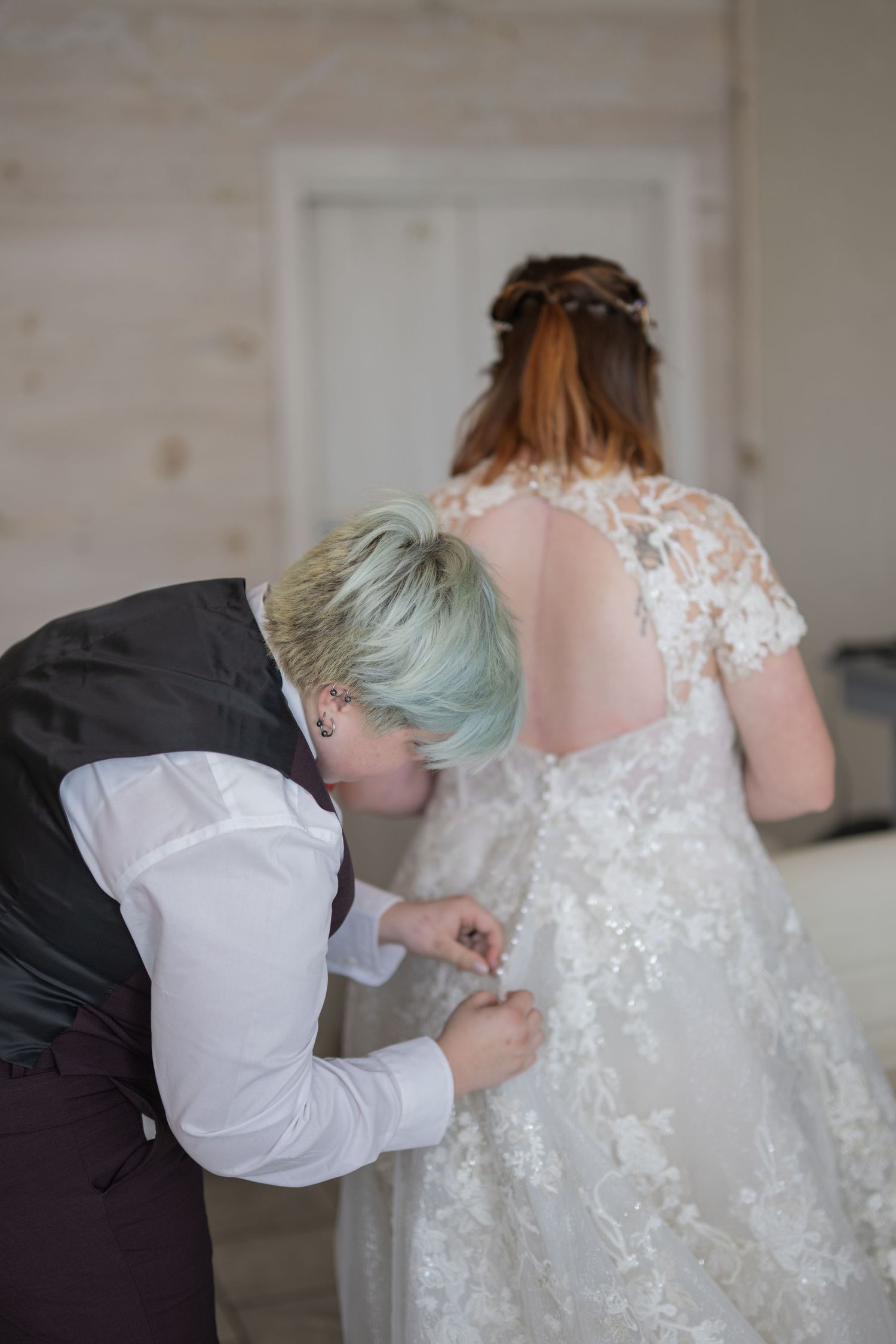 Person with short teal hair buttoning the back of a lace wedding dress on a bride with red hair.