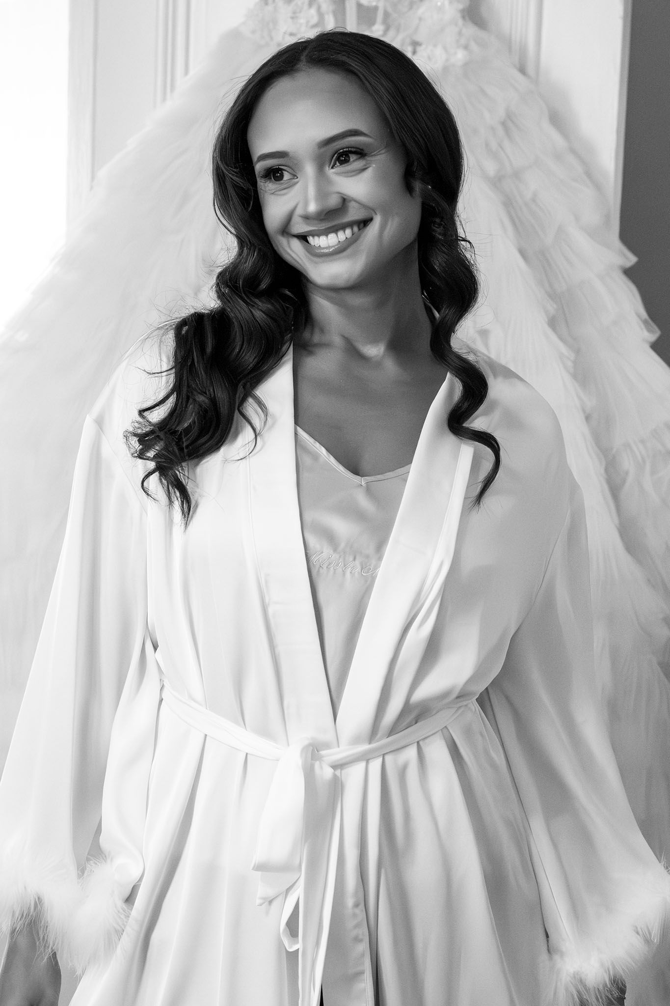 Smiling bride in white satin robe with feather cuffs poses in front of a wedding dress in black and white photo.