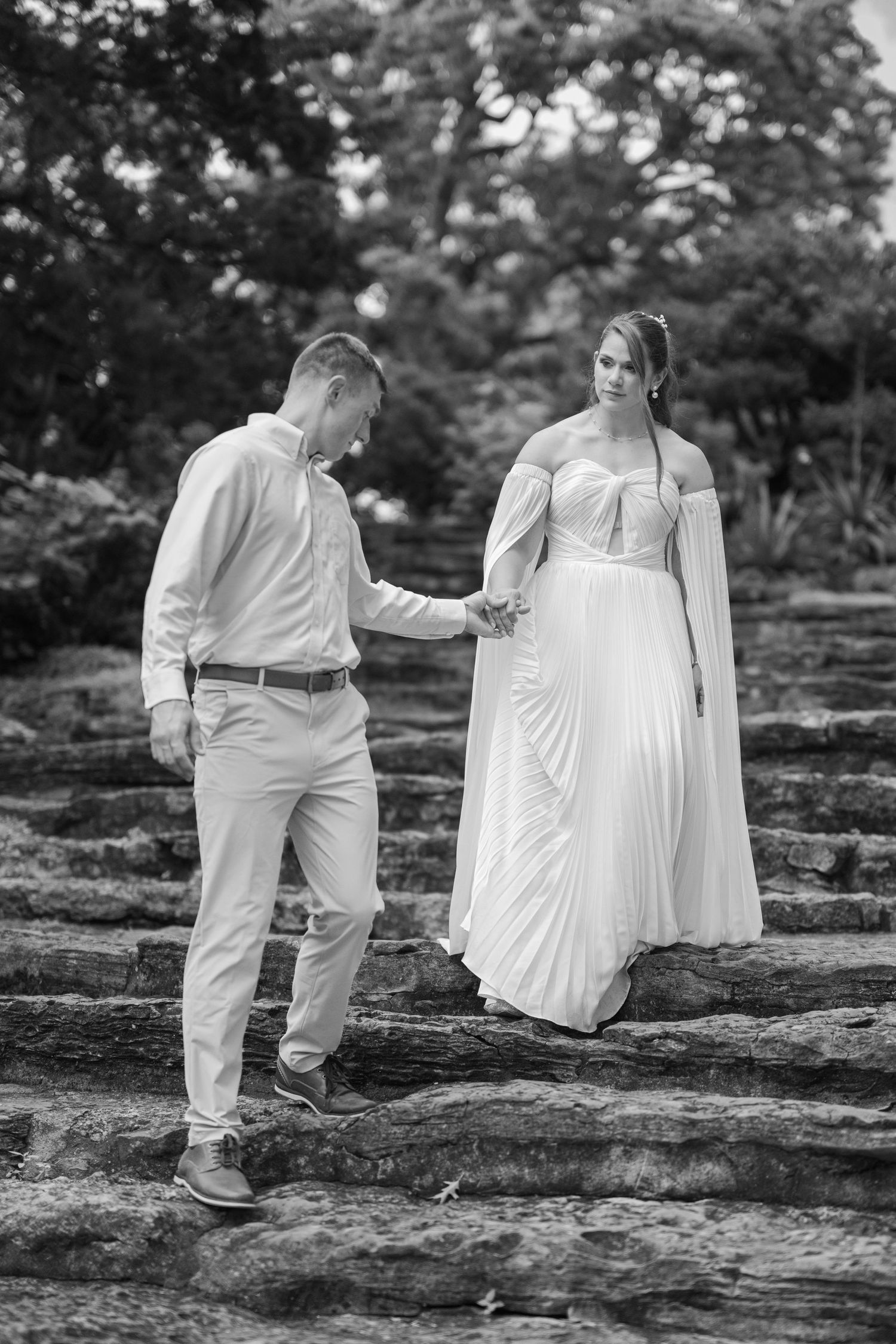 Black and white photo of a bride and groom holding hands while walking down stone steps in a lush garden.