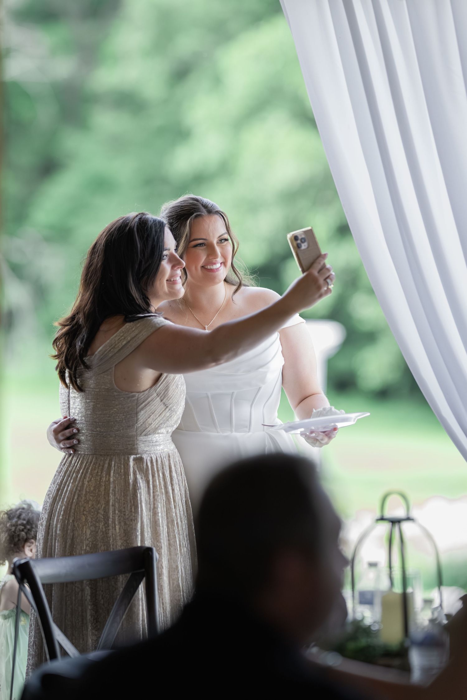 Bride and bridesmaid taking a selfie together at wedding reception near white curtains.
