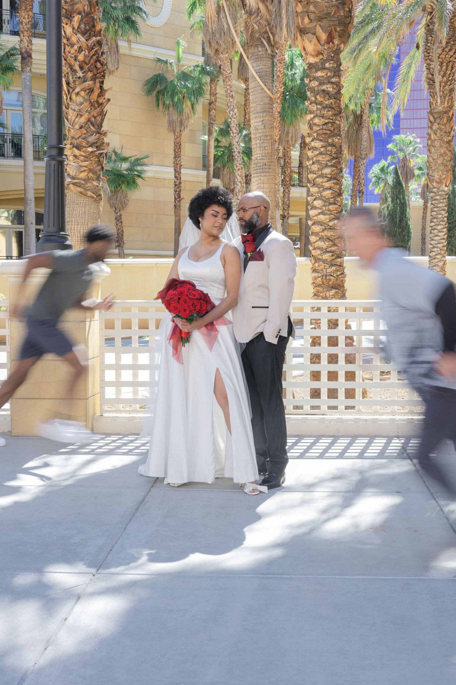 Bride and groom pose with red bouquet outdoors among palm trees, with blurred passersby in motion around them.