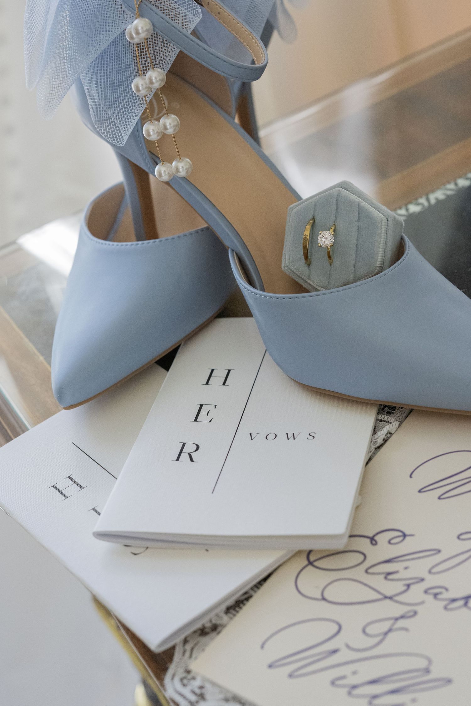 Light blue wedding heels with pearl details rest on HER vows booklets beside a diamond engagement ring in a velvet box.