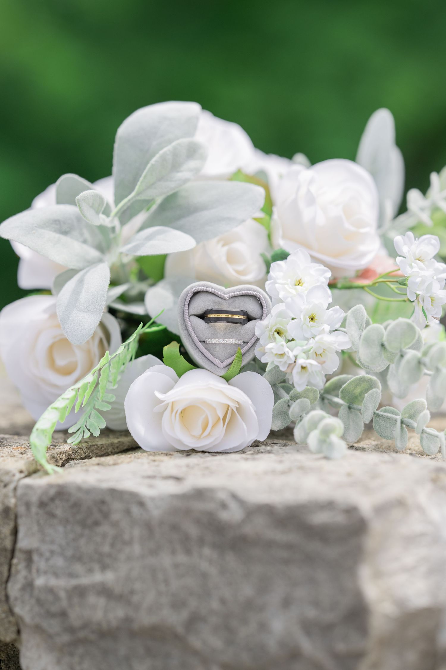 Wedding rings in heart-shaped box nestled among white roses and greenery on stone surface.