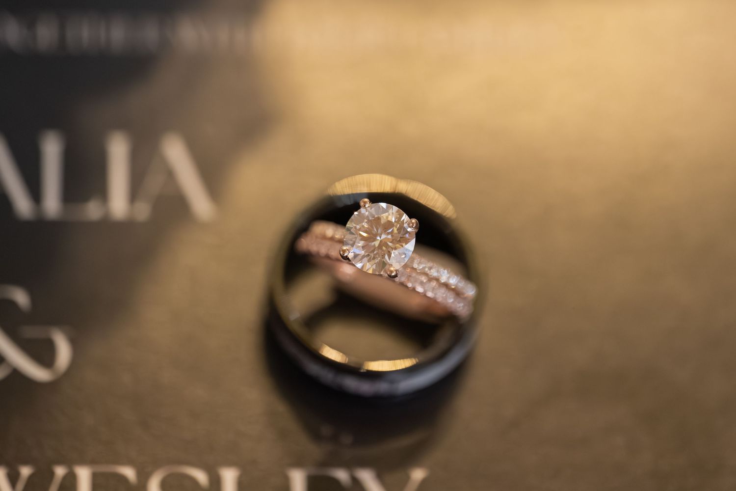 Three wedding rings stacked together on an invitation card bearing the names Alia and Wesley, featuring a diamond engagement ring.