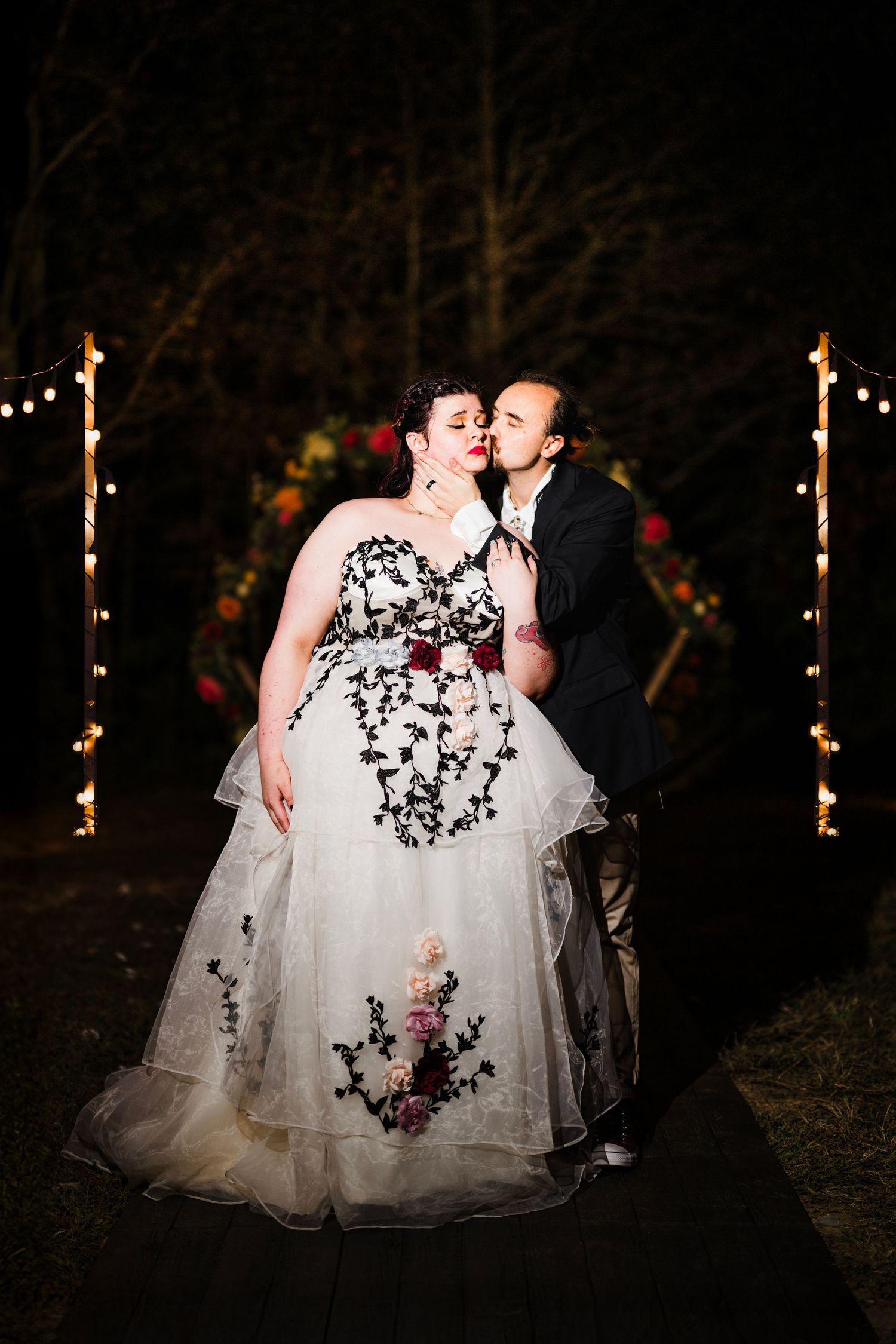 Bride in black floral gown and groom in dark suit share a kiss outdoors at night, framed by string lights.