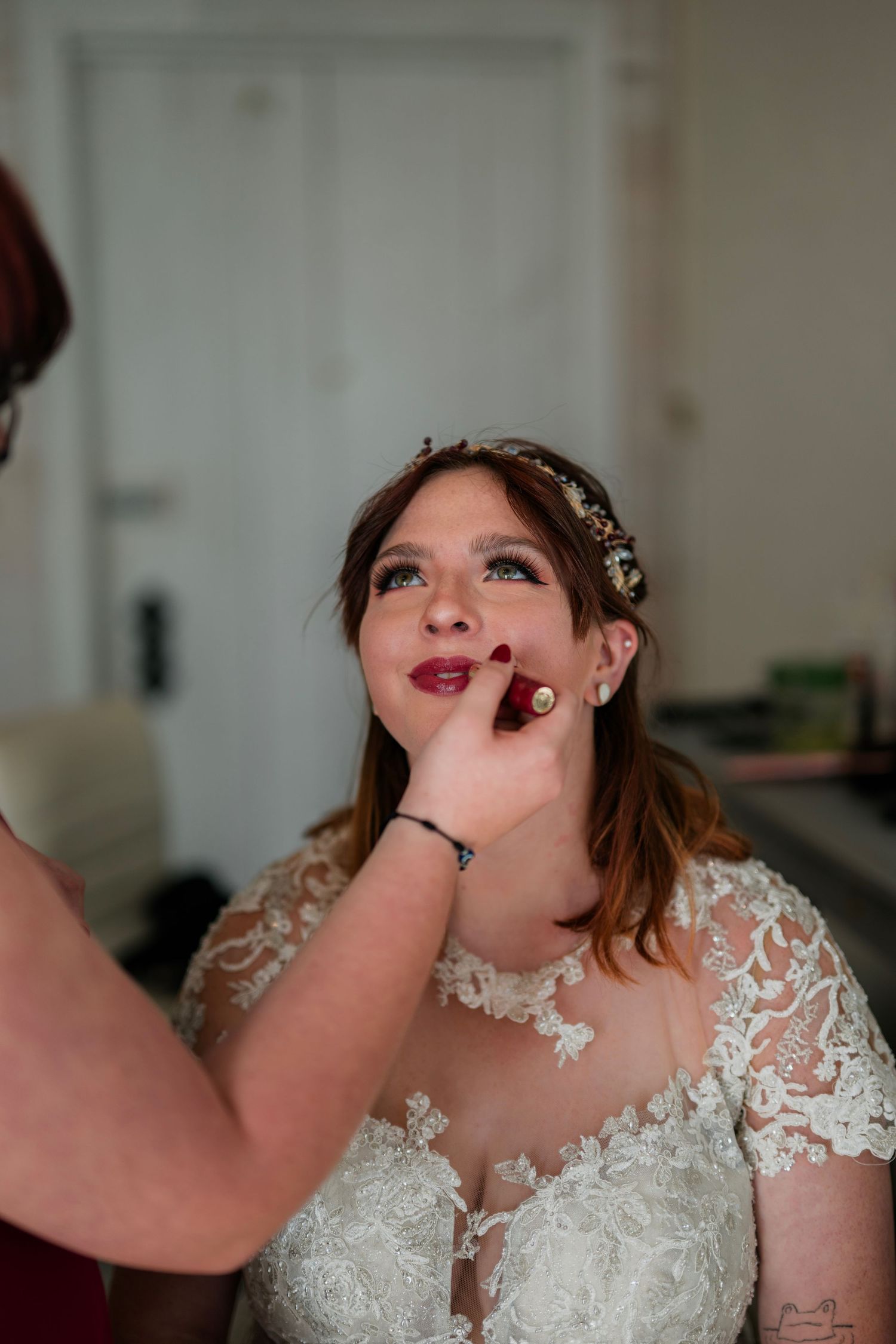Bride in lace wedding dress getting red lipstick applied by makeup artist during bridal preparation.